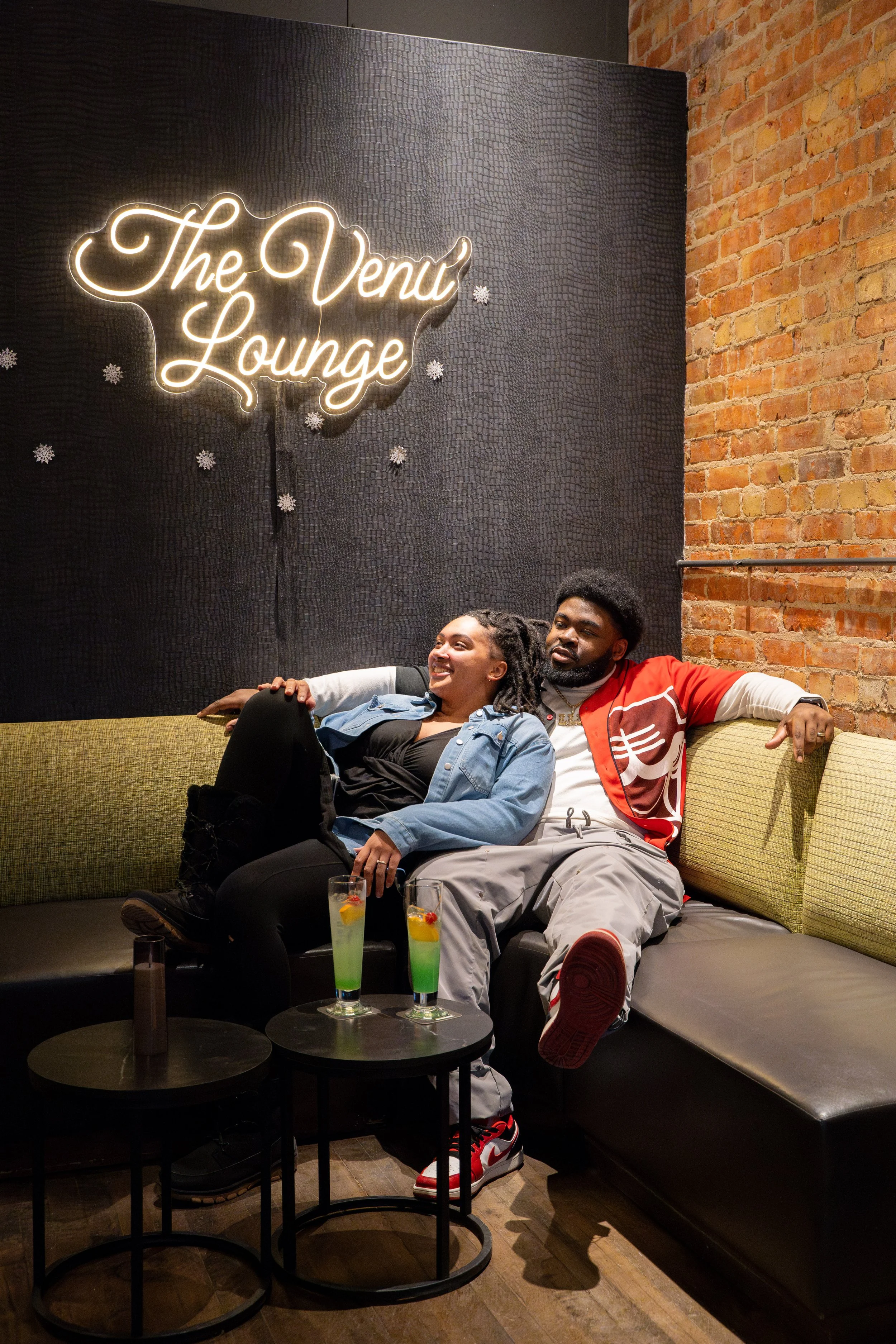 A smiling couple sitting on a green and black corner sofa in The Venu Lounge. The woman is leaning on the man, who has his arm around her. There are two colorful cocktails on a black round table in front of them. The background features a black wall 