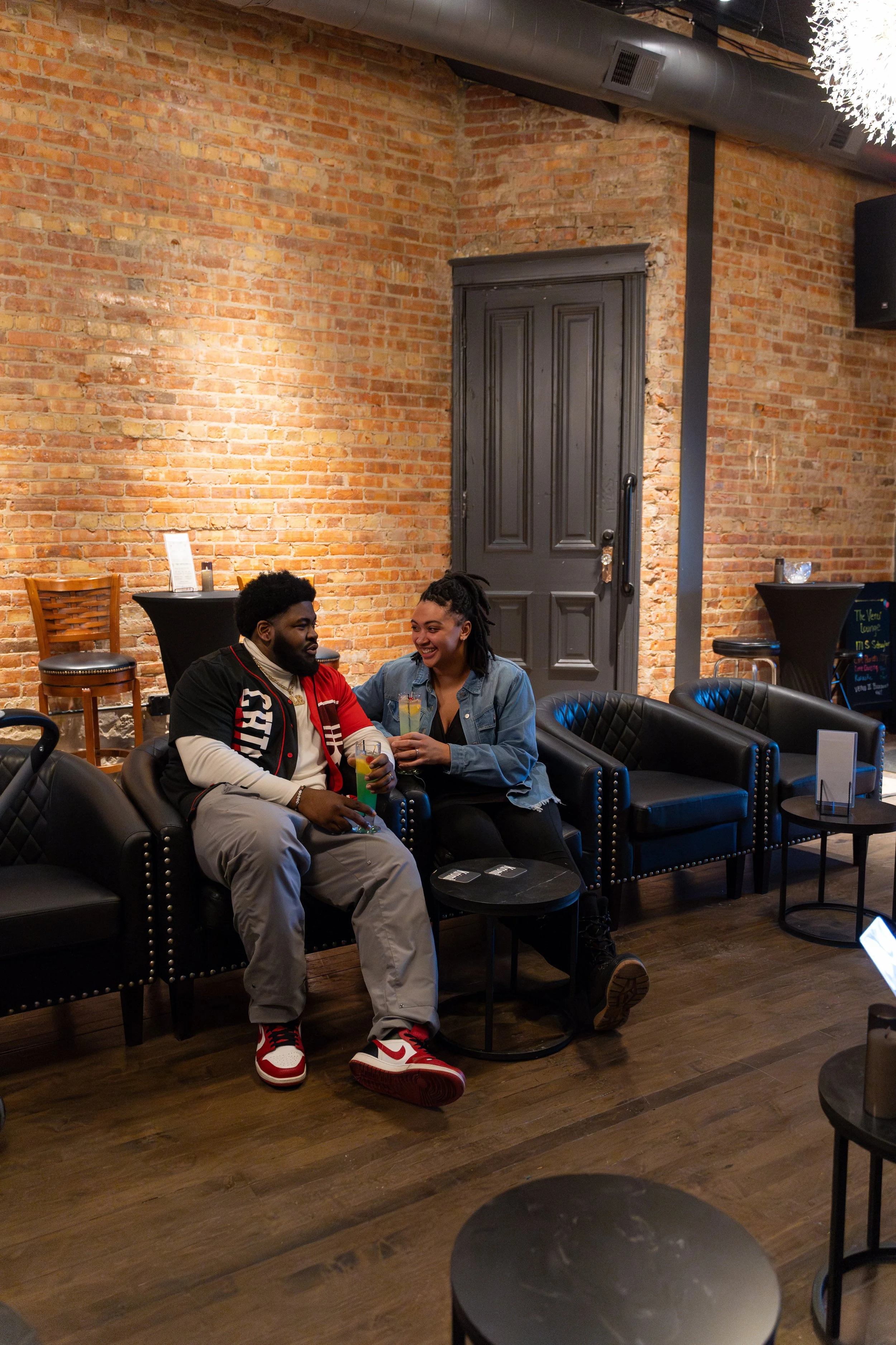 Two people sitting on a black leather couch in a bar or lounge, smiling and holding drinks, with a brick wall backdrop.