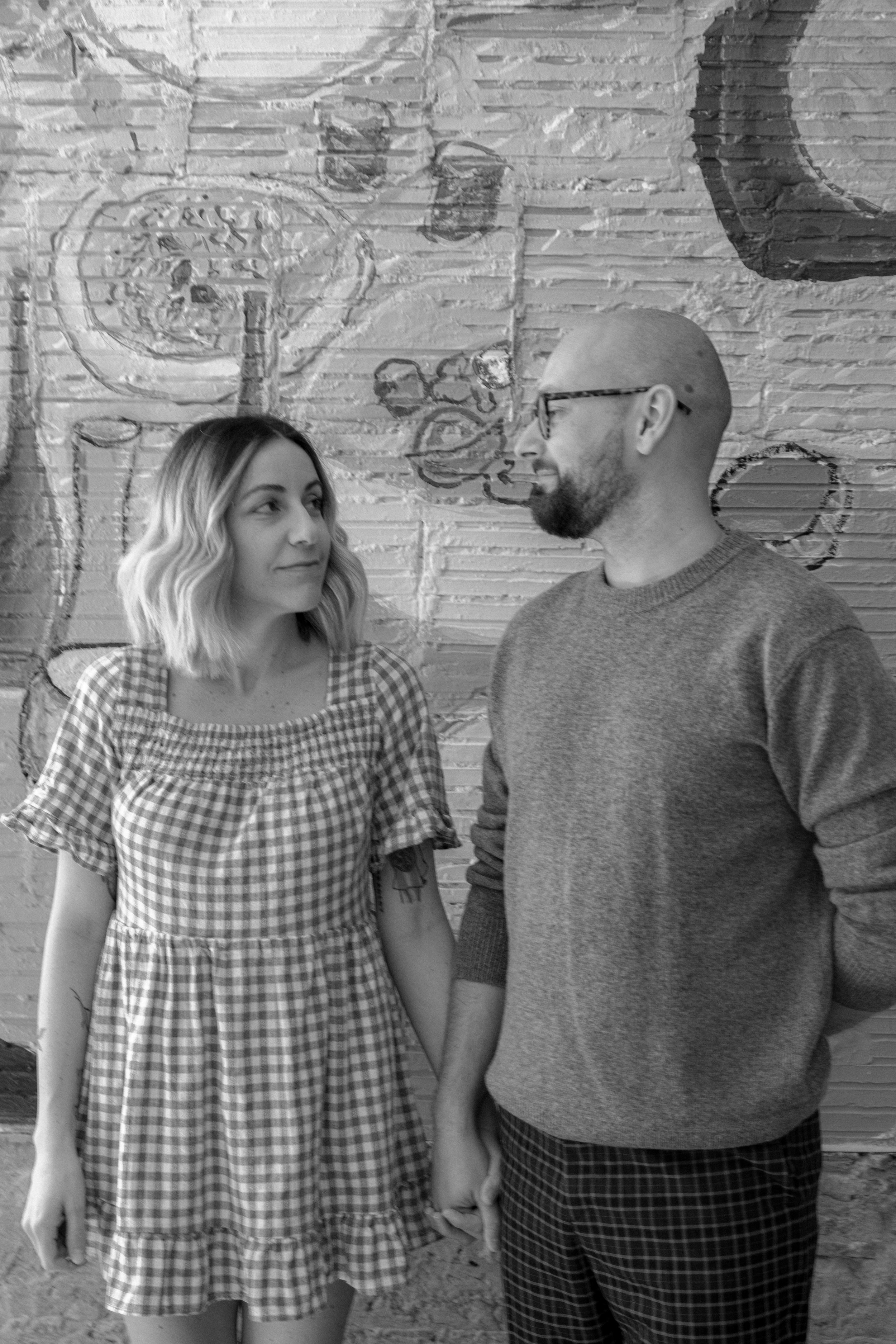 A woman and a man standing hand in hand in front of a brick wall with art, looking at each other.