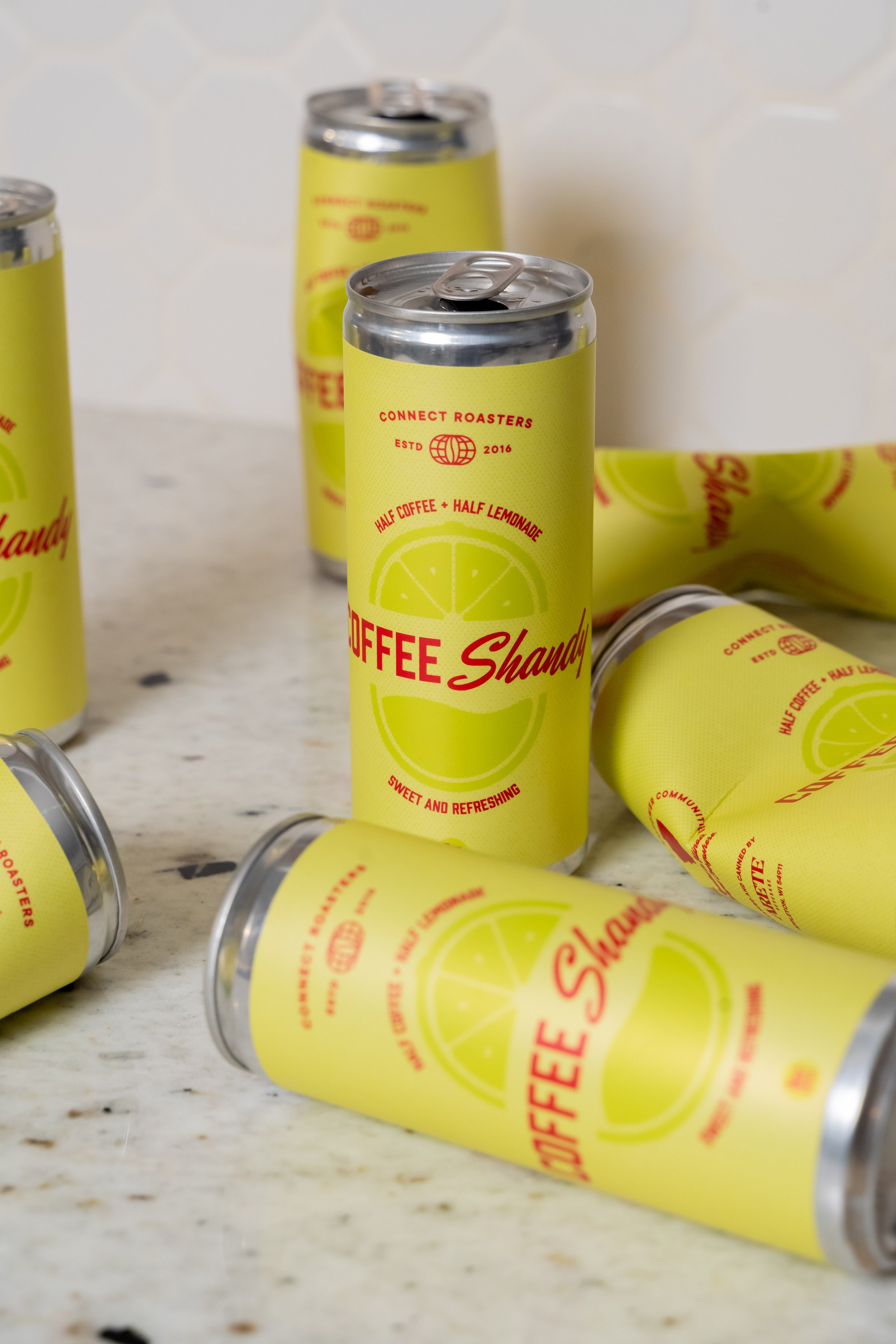 Several cans of lemonade with yellow labels and red text, some standing upright and others lying down, on a white countertop with a tile wall background.
