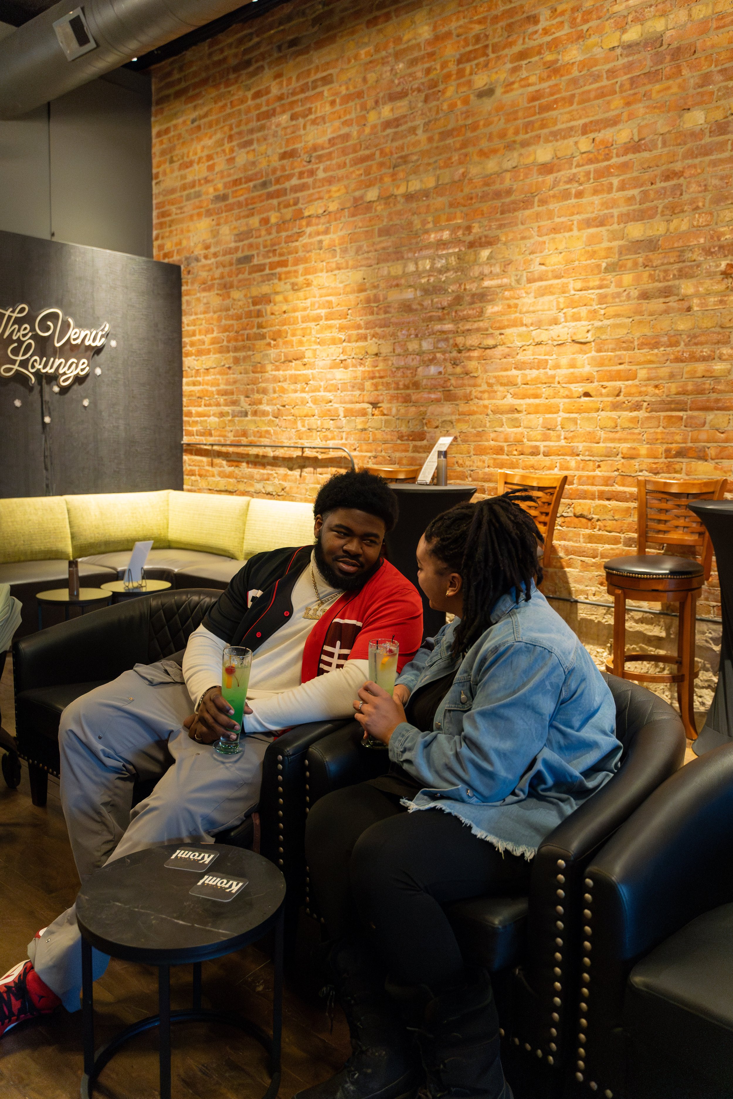 Two people sitting on black leather chairs in a lounge, holding colorful drinks and talking, with exposed brick wall and bar stools in the background.
