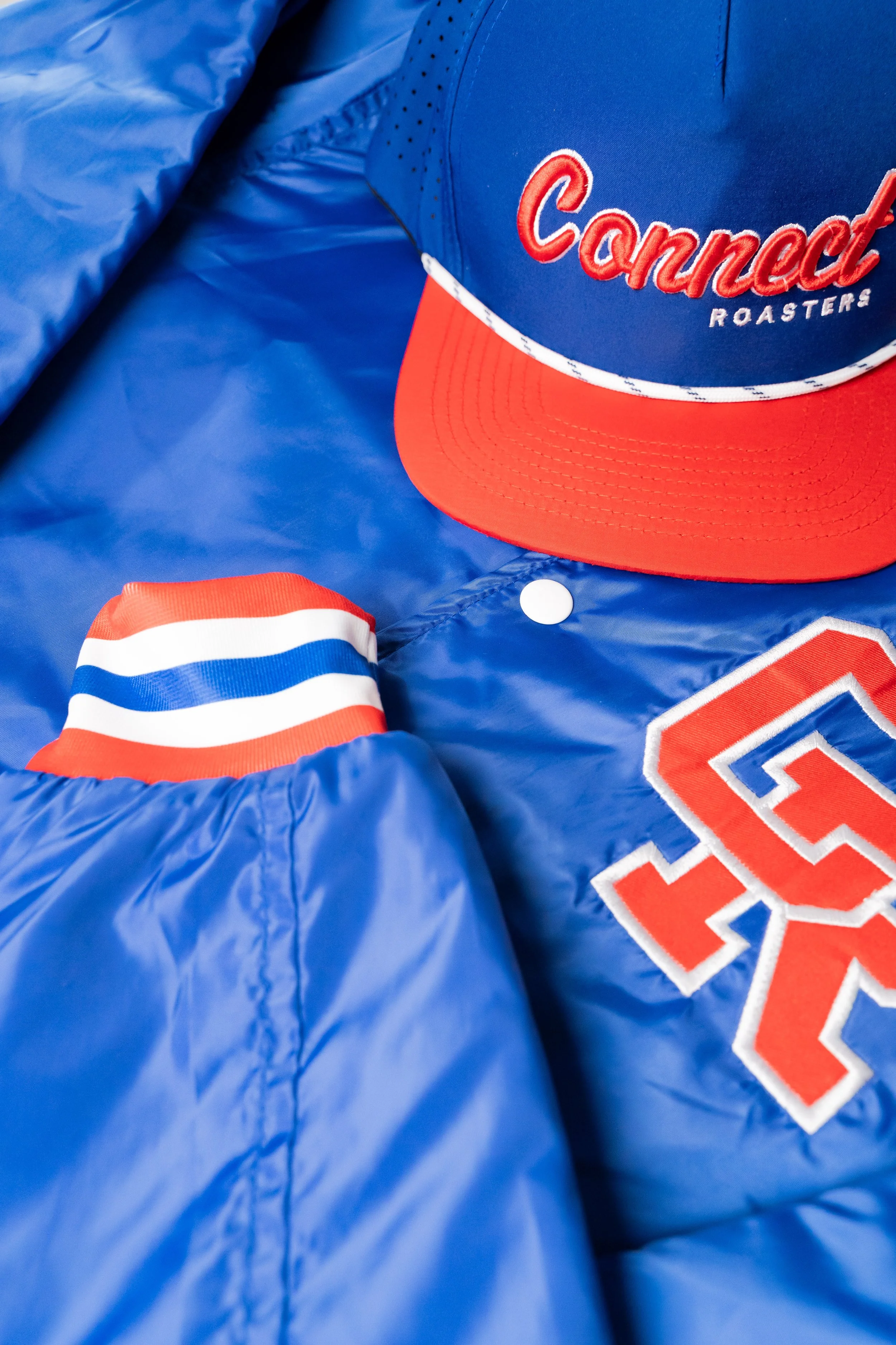 A blue jacket with red, white, and blue striped cuff, a baseball cap with red bill and blue crown embroidered with 'Connect Roasters', and a partial embroidered word 'USA' on the jacket.