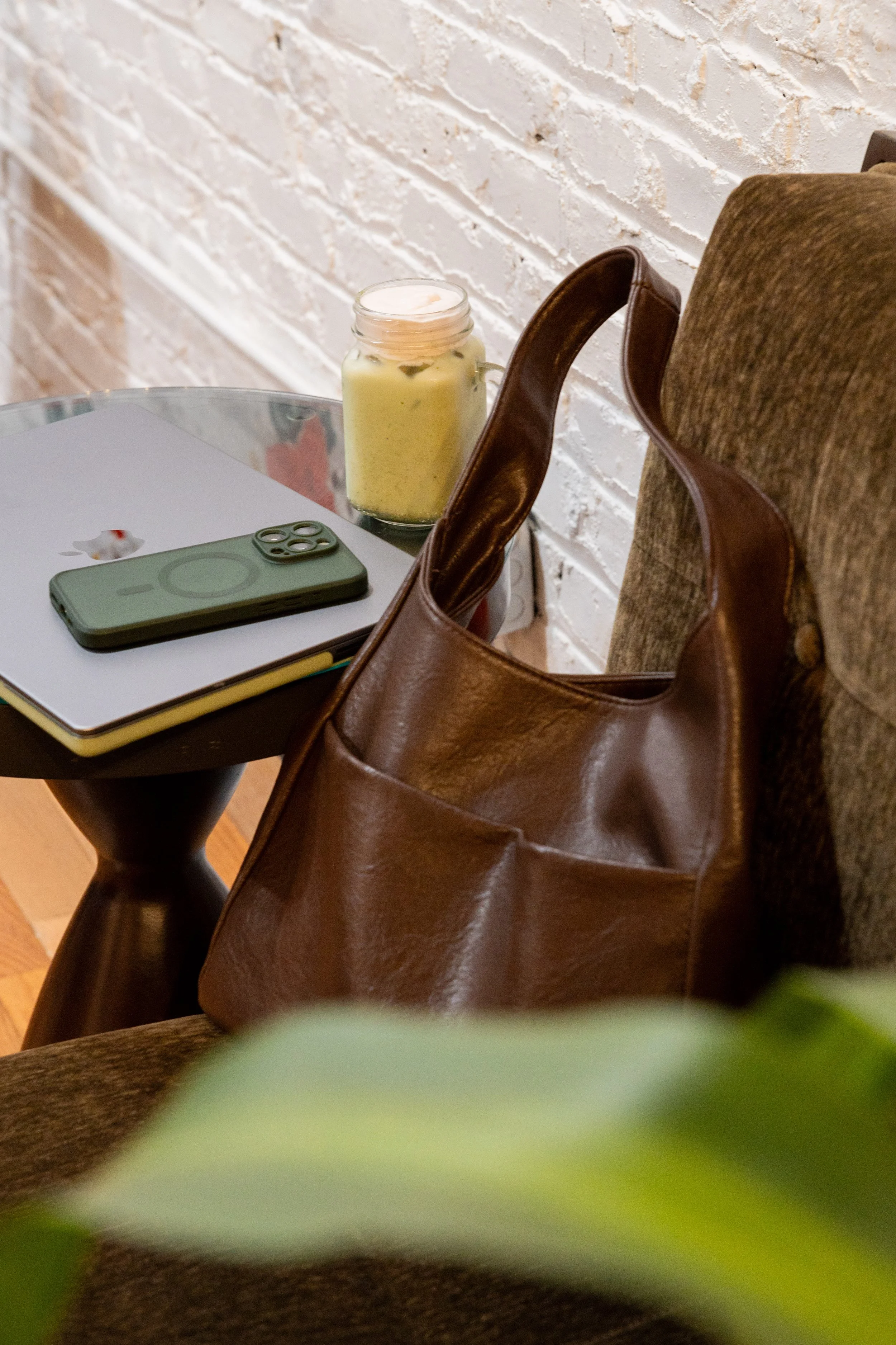 A brown leather handbag hanging on the arm of a brown upholstered chair, a Starbucks logo on a closed notebook, a smartphone on top of the notebook, a jar candle, and part of a green plant in the foreground, on a round table against a white brick wal