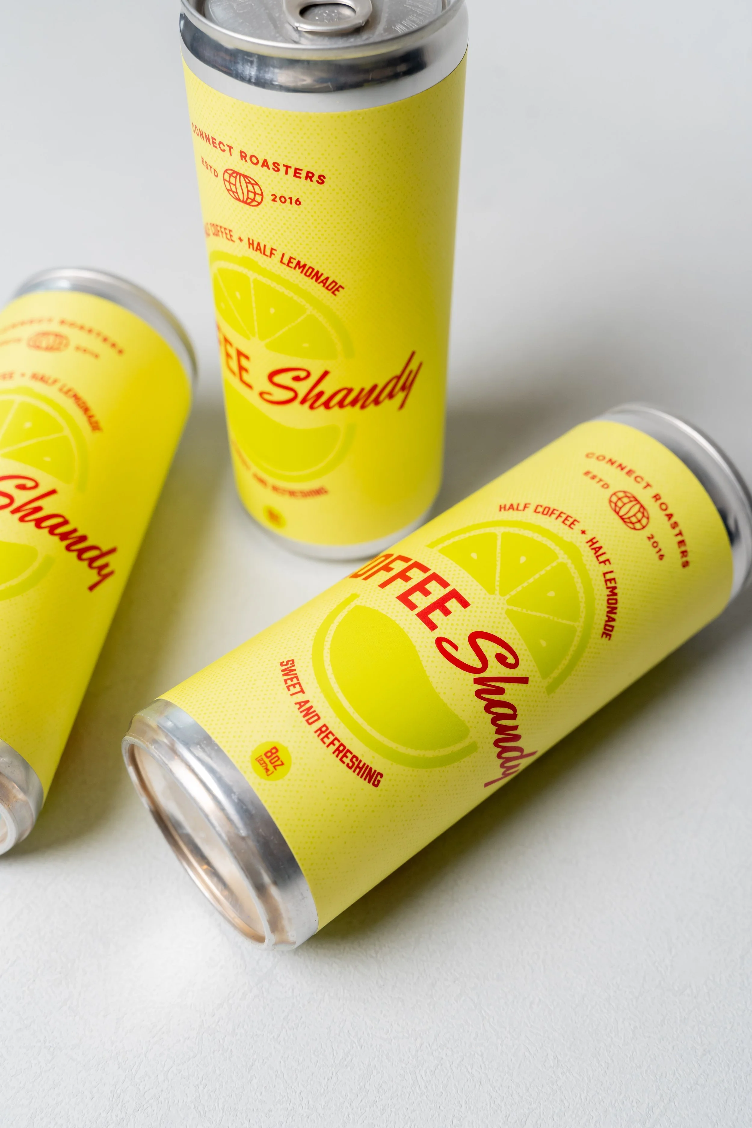 Three aluminum cans with yellow labels featuring lemon illustrations. The cans are labeled "Coffee Shandy" with text indicating half coffee and half lemonade, and phrases like "sweet and refreshing."