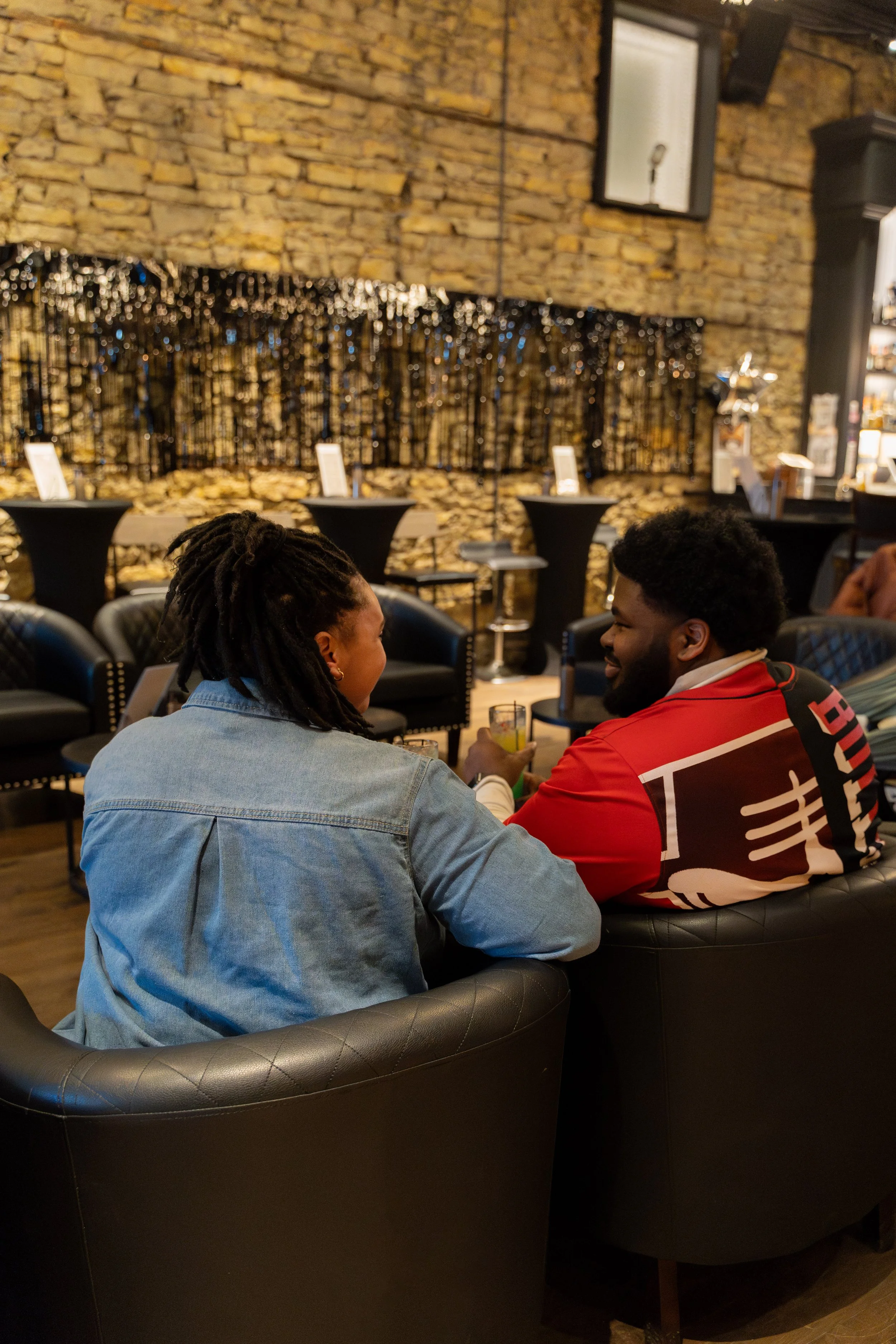 Two people sitting on a black leather couch in a bar or lounge, having a conversation and sharing a drink, with a brick wall and string lights in the background.