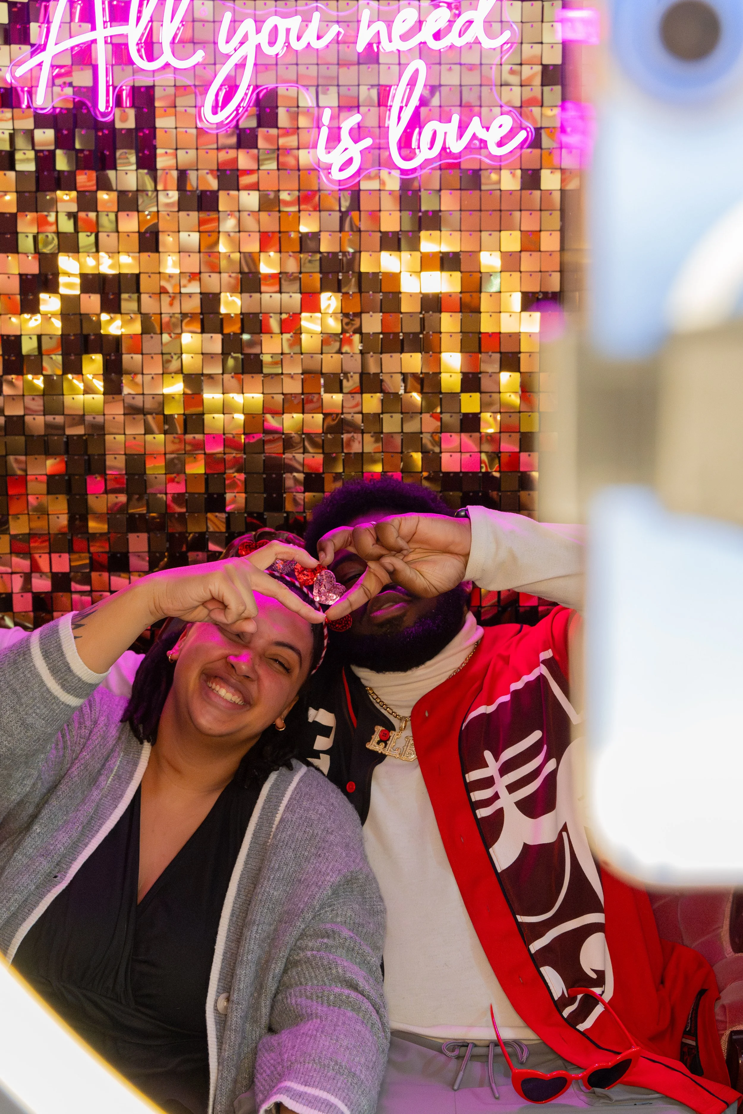 Two people smiling and posing together making a heart shape with their hands in front of a glittery gold mosaic background with pink neon sign that reads 'All you need is love'.