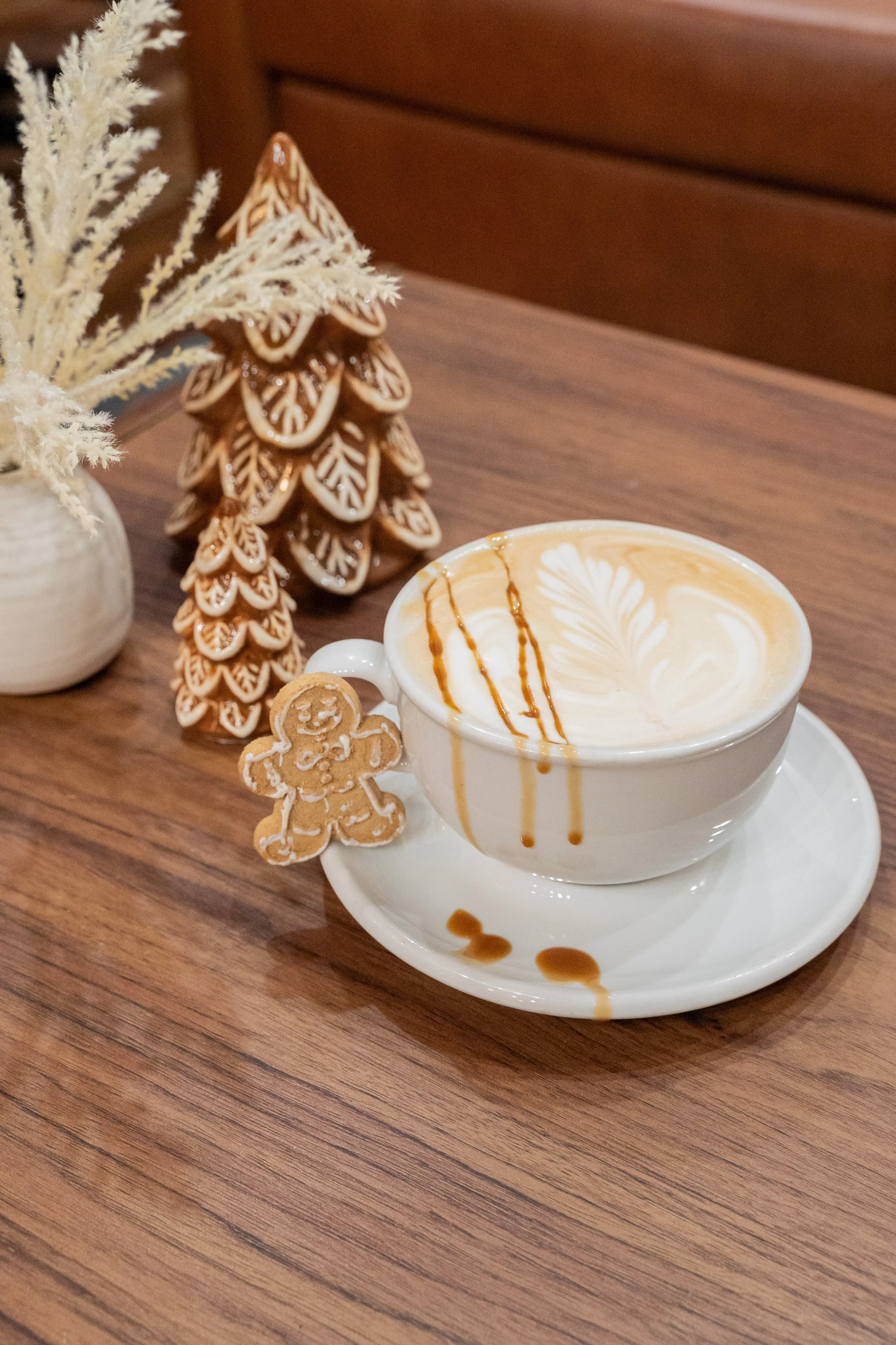 A cup of cappuccino with latte art and caramel drizzle on top, placed on a saucer with caramel drops, alongside gingerbread cookies, a decorative gingerbread tree, and a small vase with artificial snow-covered branches on a wooden table.
