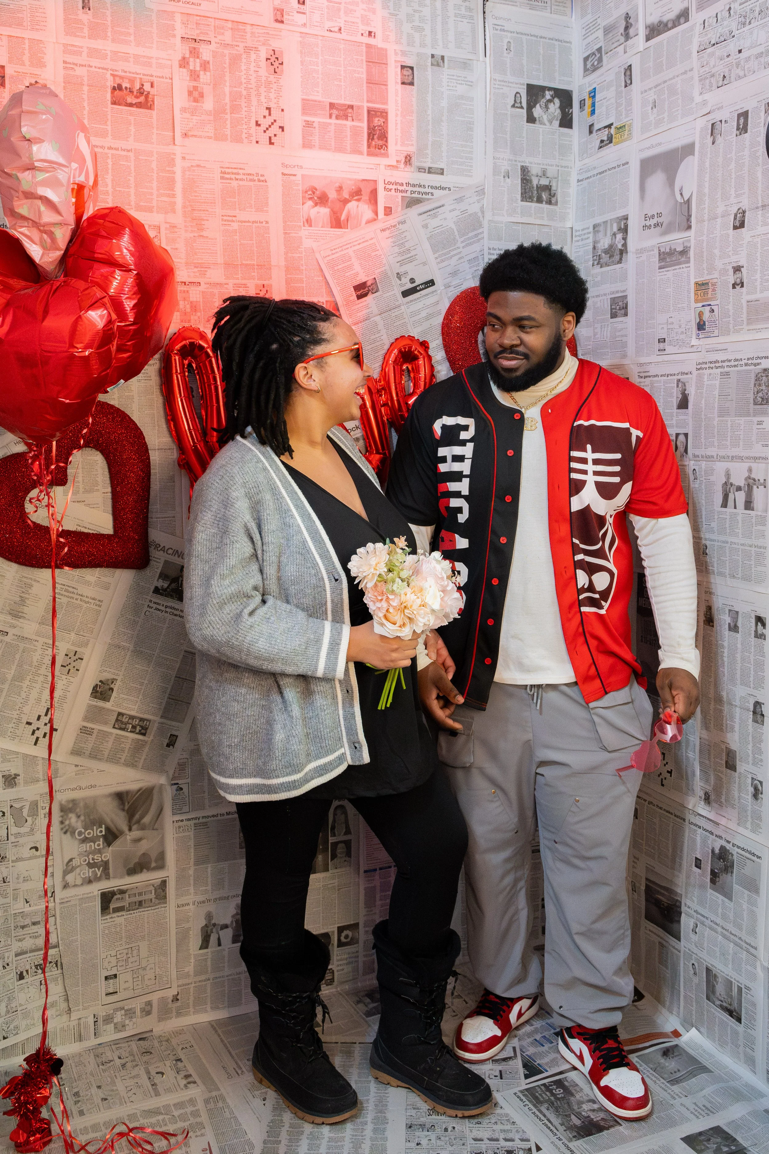 A couple standing close together inside a decorated room with newspaper-covered walls, holding a bouquet and Valentine's Day balloons, celebrating Valentine's Day.