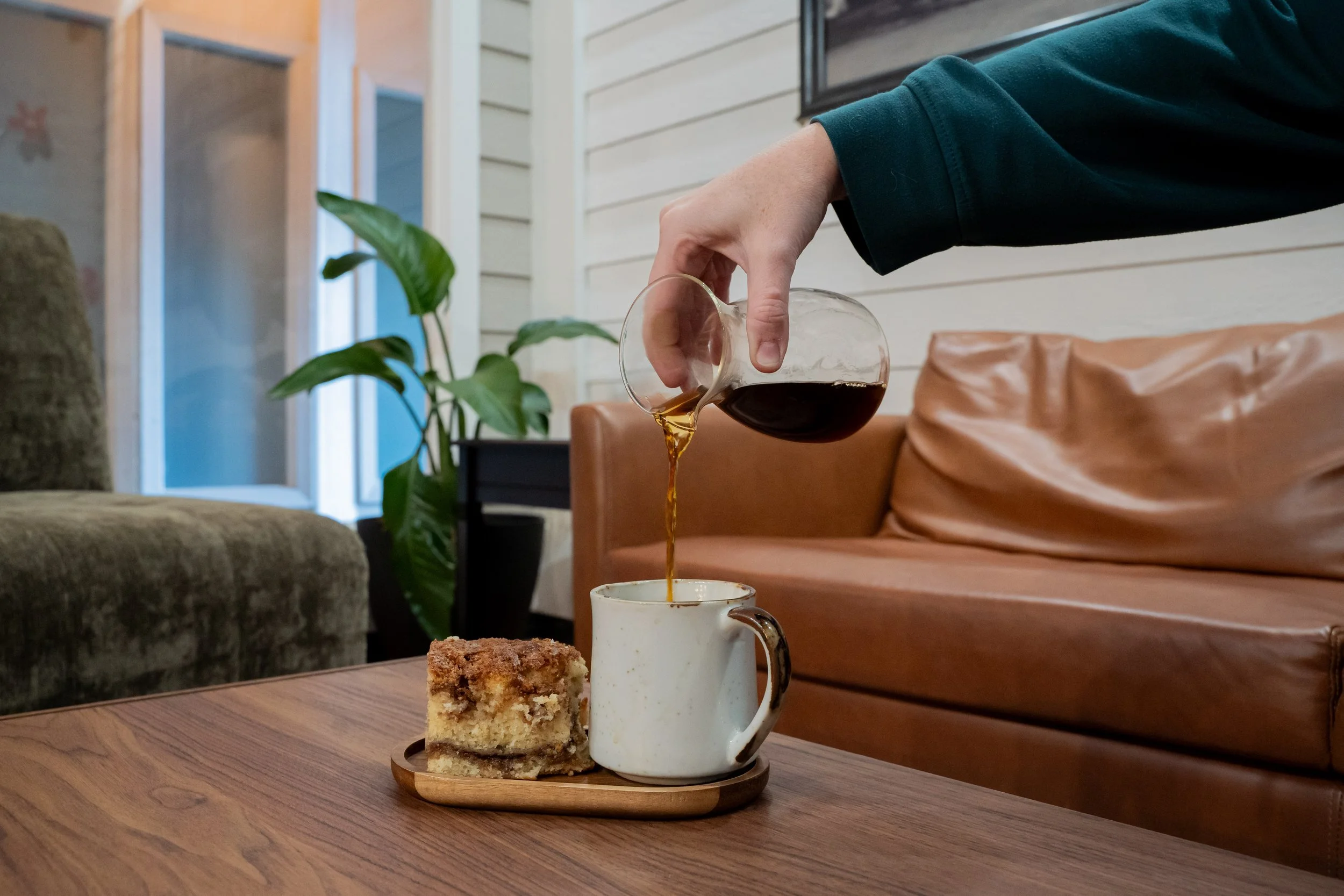 A person pours coffee into a white mug on a wooden table, with a slice of crumb cake on a tray nearby, in a cozy indoor setting with sofas and a plant in the background.