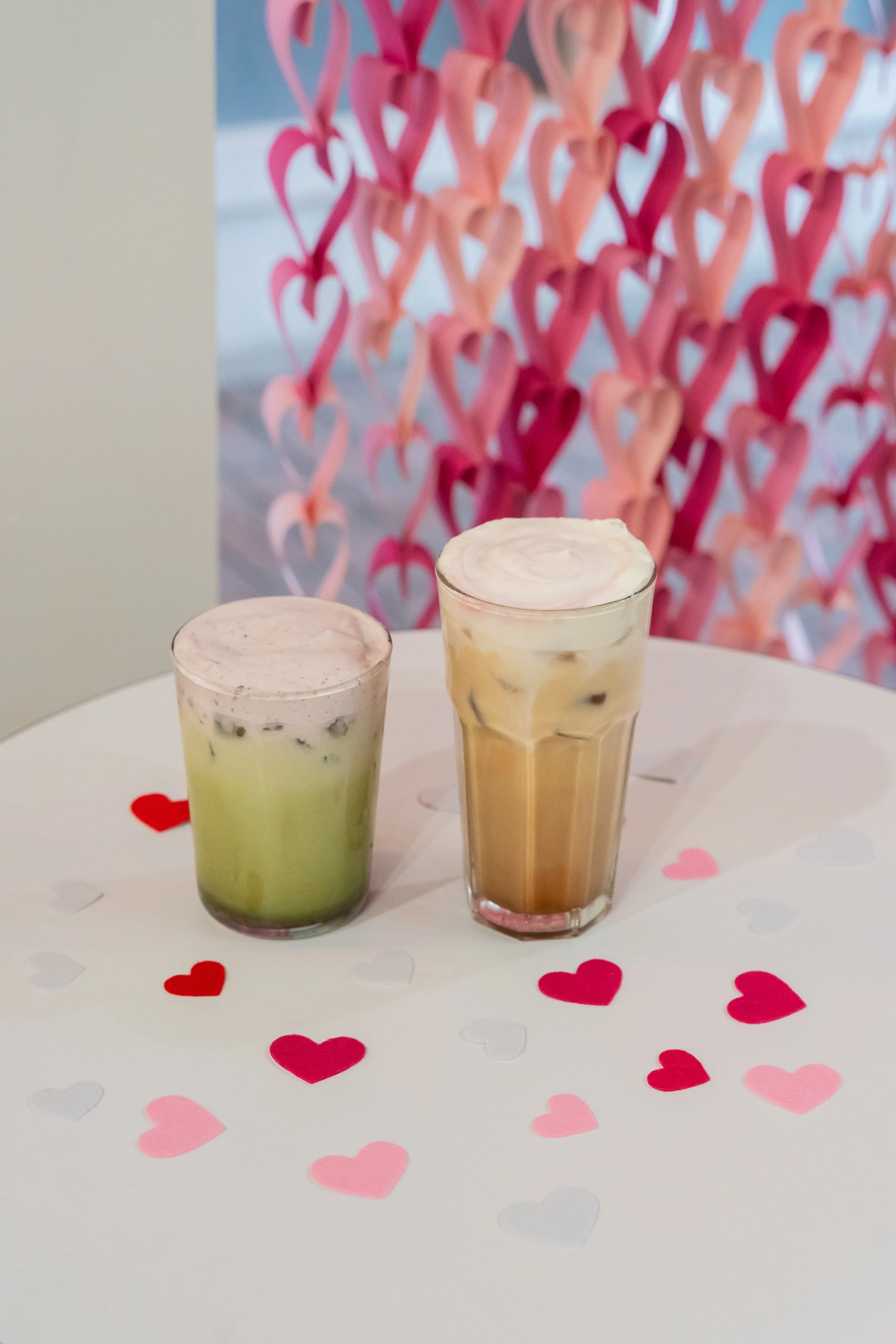 Two iced coffee drinks, one with green tea and black tapioca pearls, the other with caramel and whipped cream, on a table decorated with red, pink, and white paper hearts, with a backdrop of pink and red paper hearts.