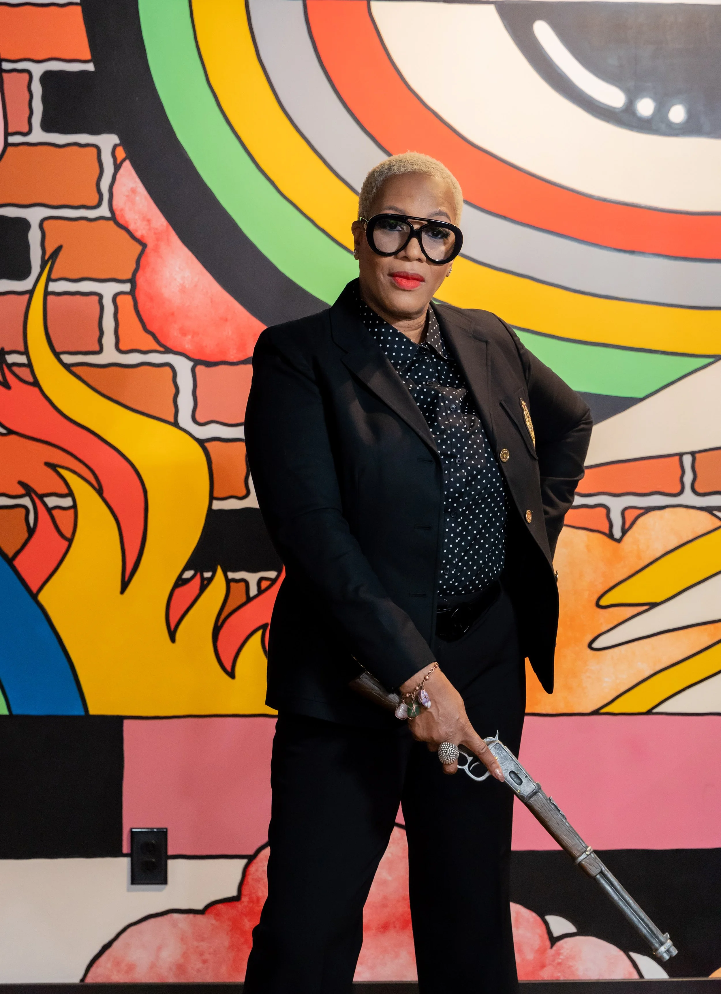 A person with short blonde hair, wearing a black suit with a polka-dot shirt, large sunglasses, and jewelry, standing in front of a colorful graffiti-style mural. The person is holding a large, vintage-style gun.