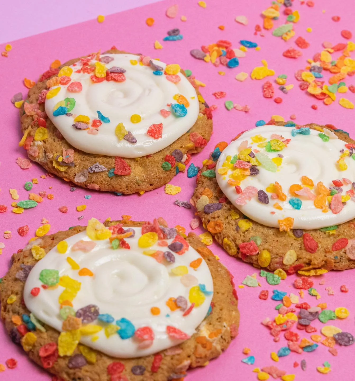 Three cookies topped with white frosting and colorful sprinkle confetti on a pink background.