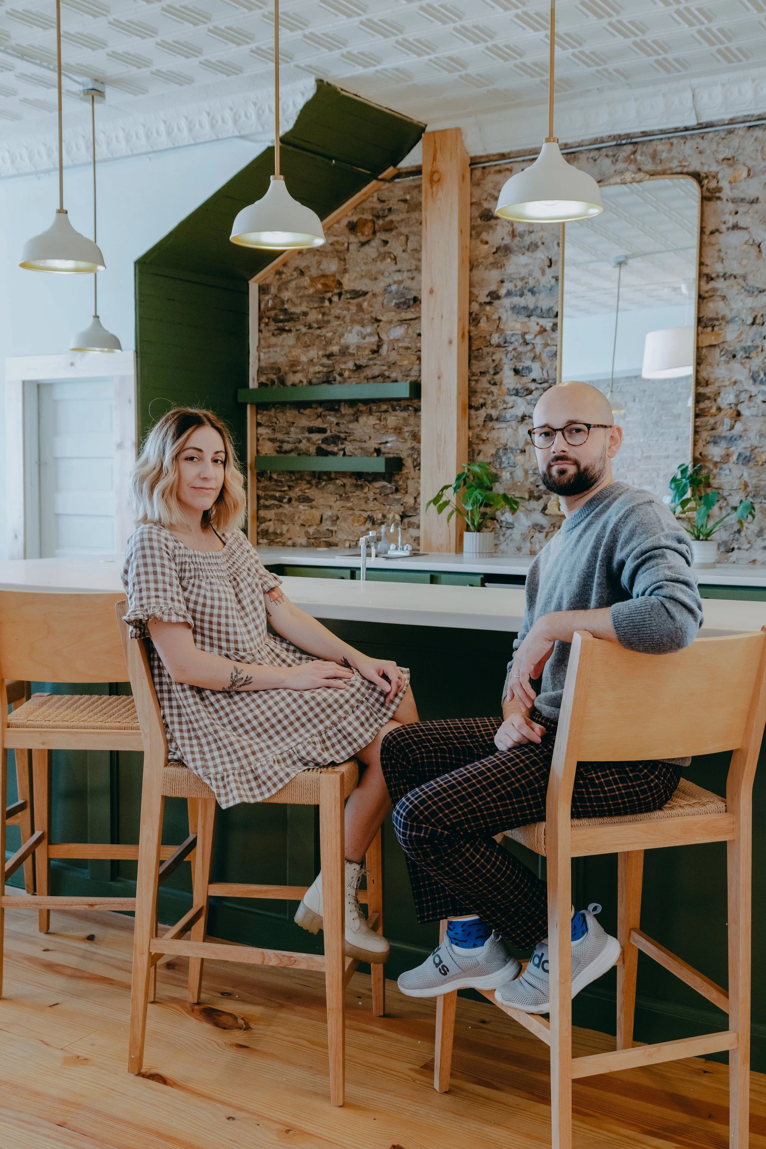 A young woman and man sitting on high wooden chairs at a kitchen island in a modern, rustic-style kitchen with exposed brick walls and hanging pendant lights.
