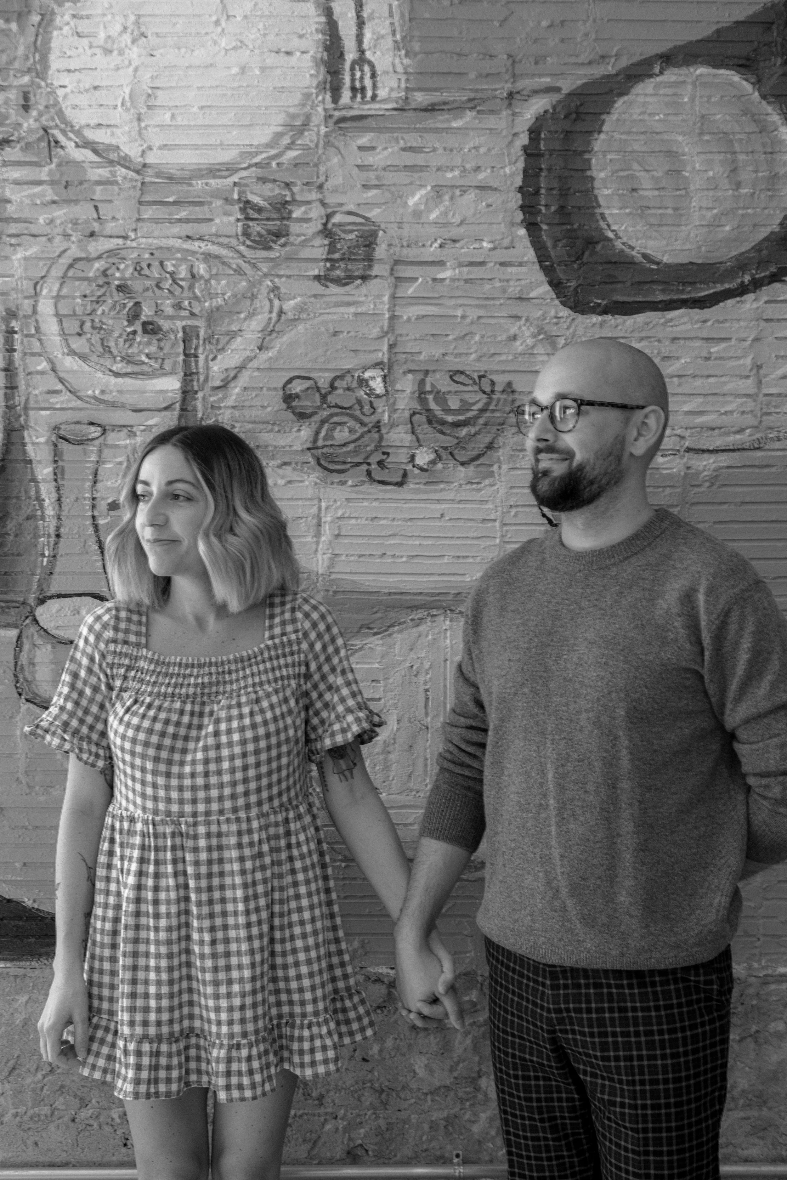 A man and woman holding hands in front of a brick wall with food-themed graffiti, including a pizza and a beverage, in a casual setting.