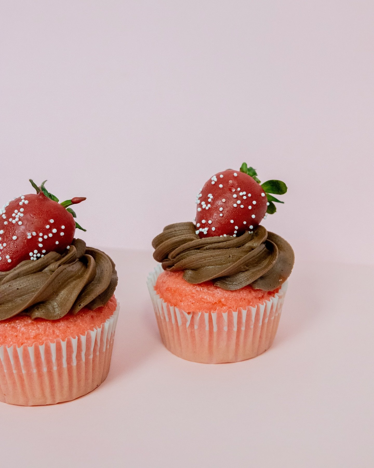 Two cupcakes with pink liners, topped with chocolate frosting and a strawberry with sprinkles, set against a light pink background.