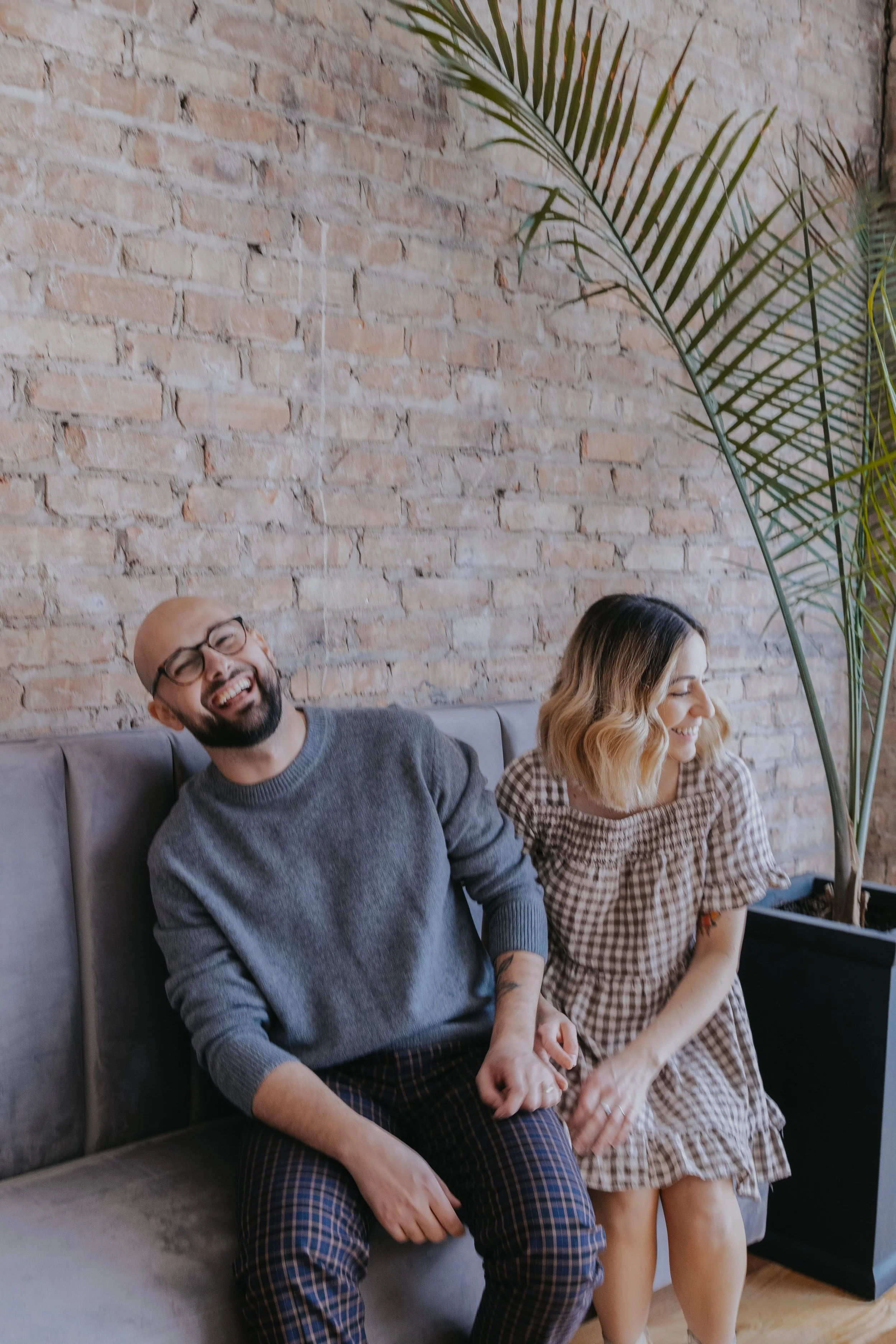A smiling man and woman sitting on a gray couch, holding hands, in front of a brick wall and a large indoor plant.