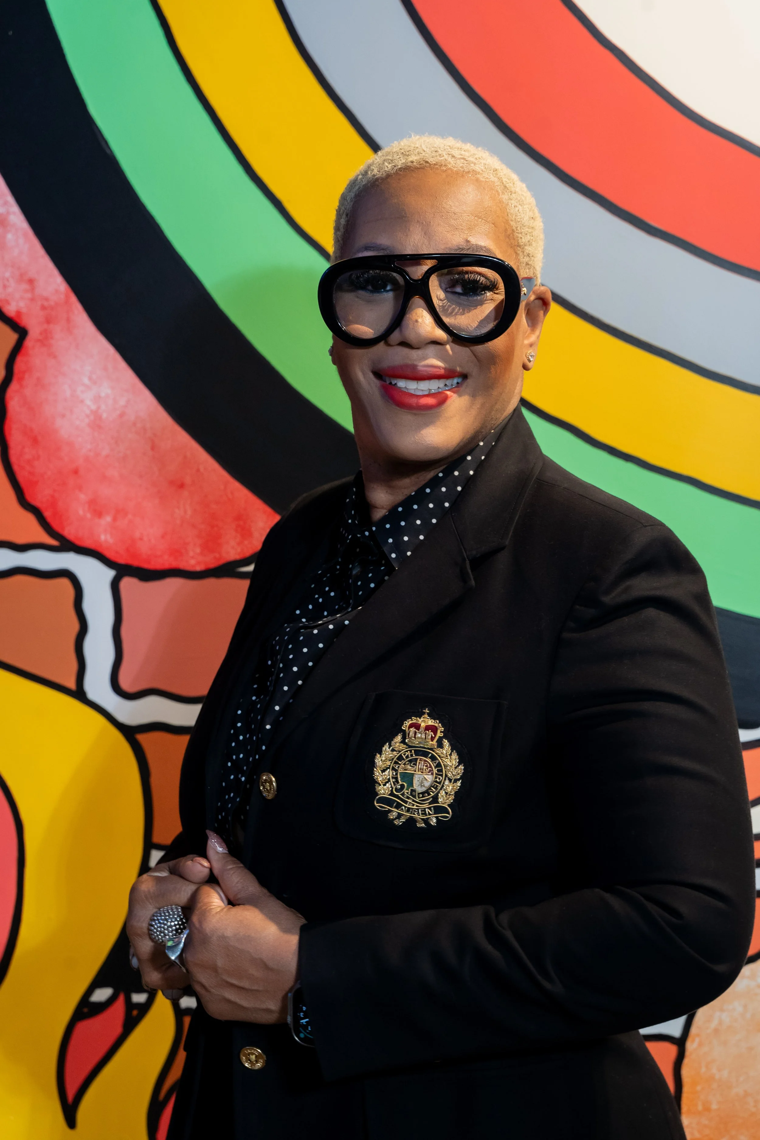 A person with short platinum blonde hair, wearing large black glasses, red lipstick, a black blazer with an embroidered crest, and a black polka dot shirt, standing in front of a colorful, abstract mural.