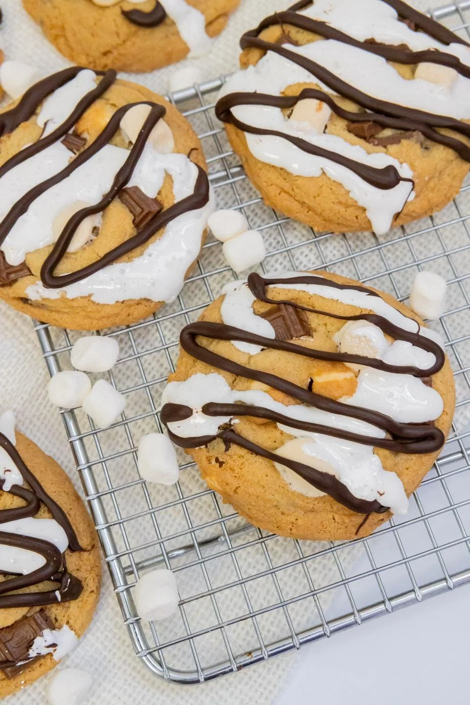 Chocolate chip cookies topped with white and dark chocolate drizzles, served on a cooling rack with mini marshmallows scattered around.