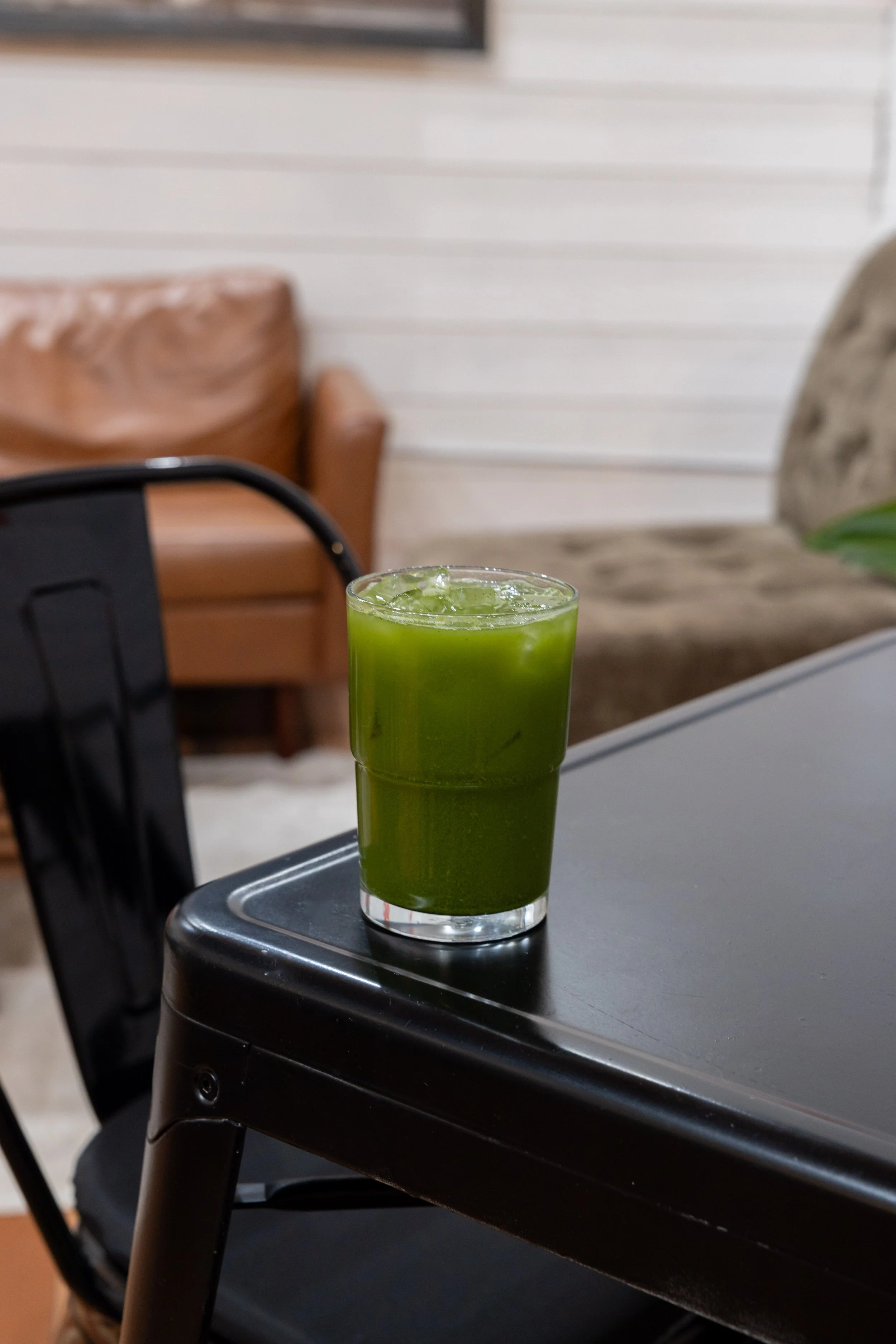 A glass of green juice with ice on a black table in a cozy indoor setting.