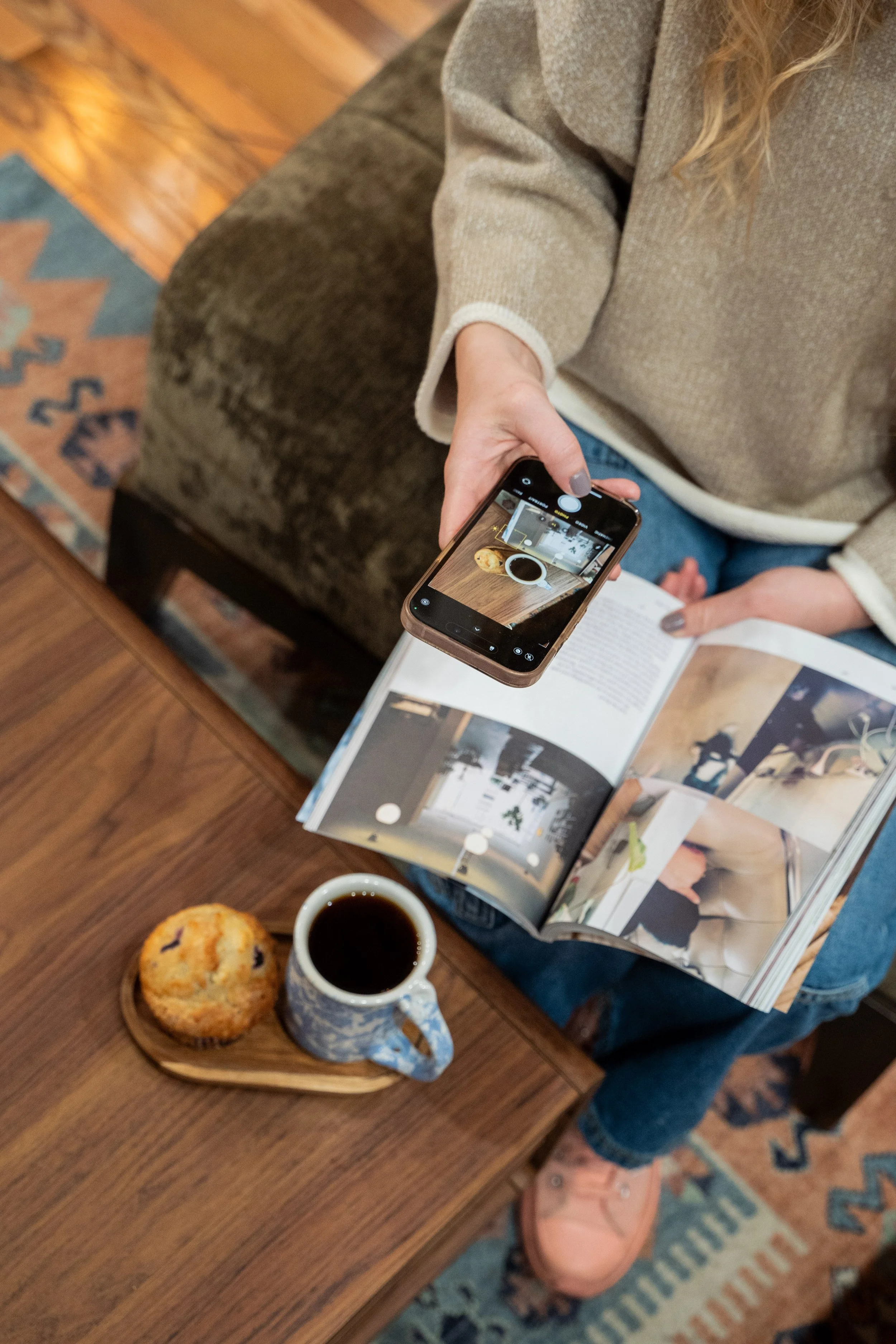 A person sitting on a couch takes a photo of a magazine with a smartphone, on a wooden table there is a cup of coffee and a muffin.