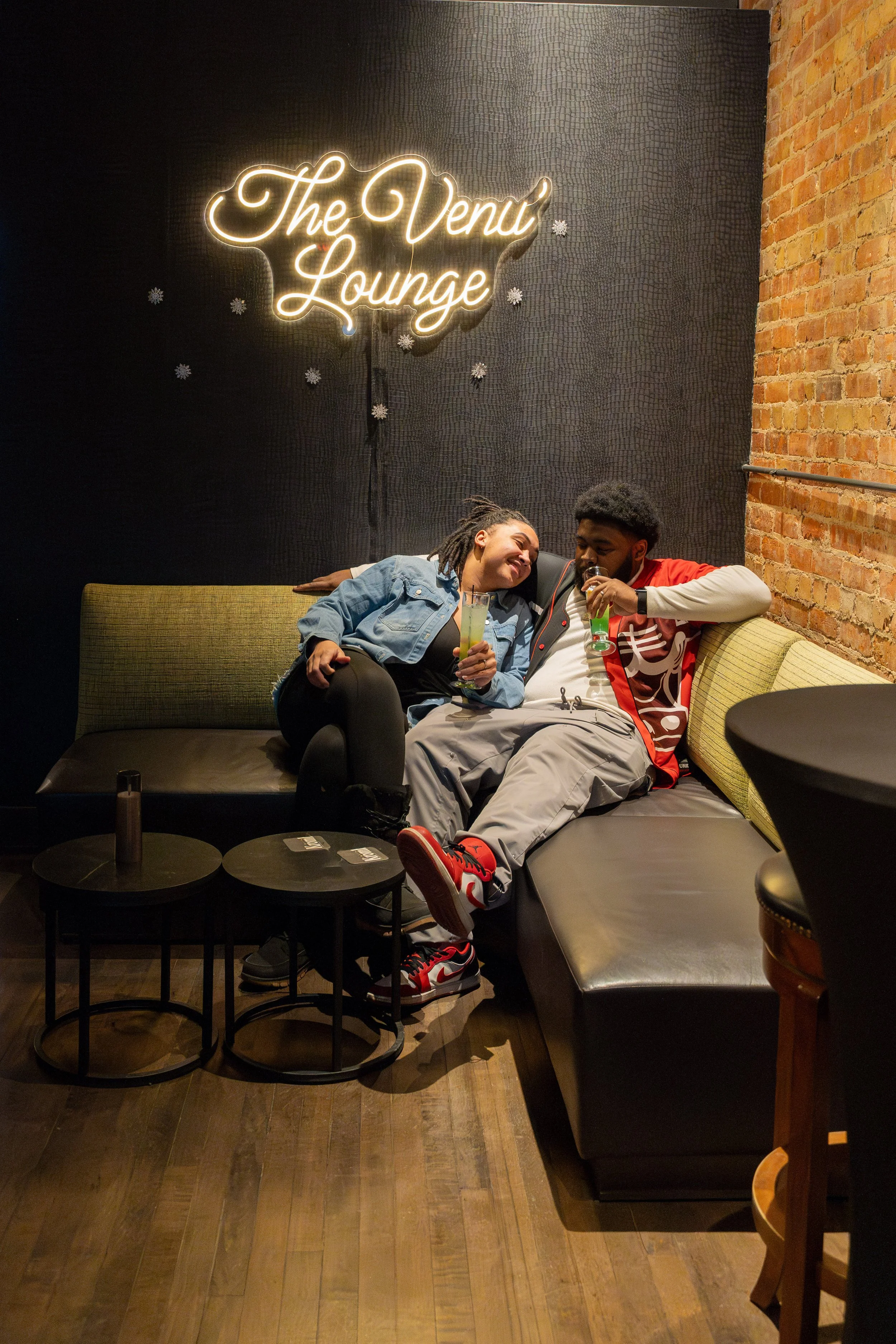 Two young adults sitting on a black couch in a lounge, smiling and enjoying drinks. The woman is leaning on the man's shoulder. Behind them, a neon sign reads 'The Venue Lounge' on a black textured wall, with a brick wall on the right side.