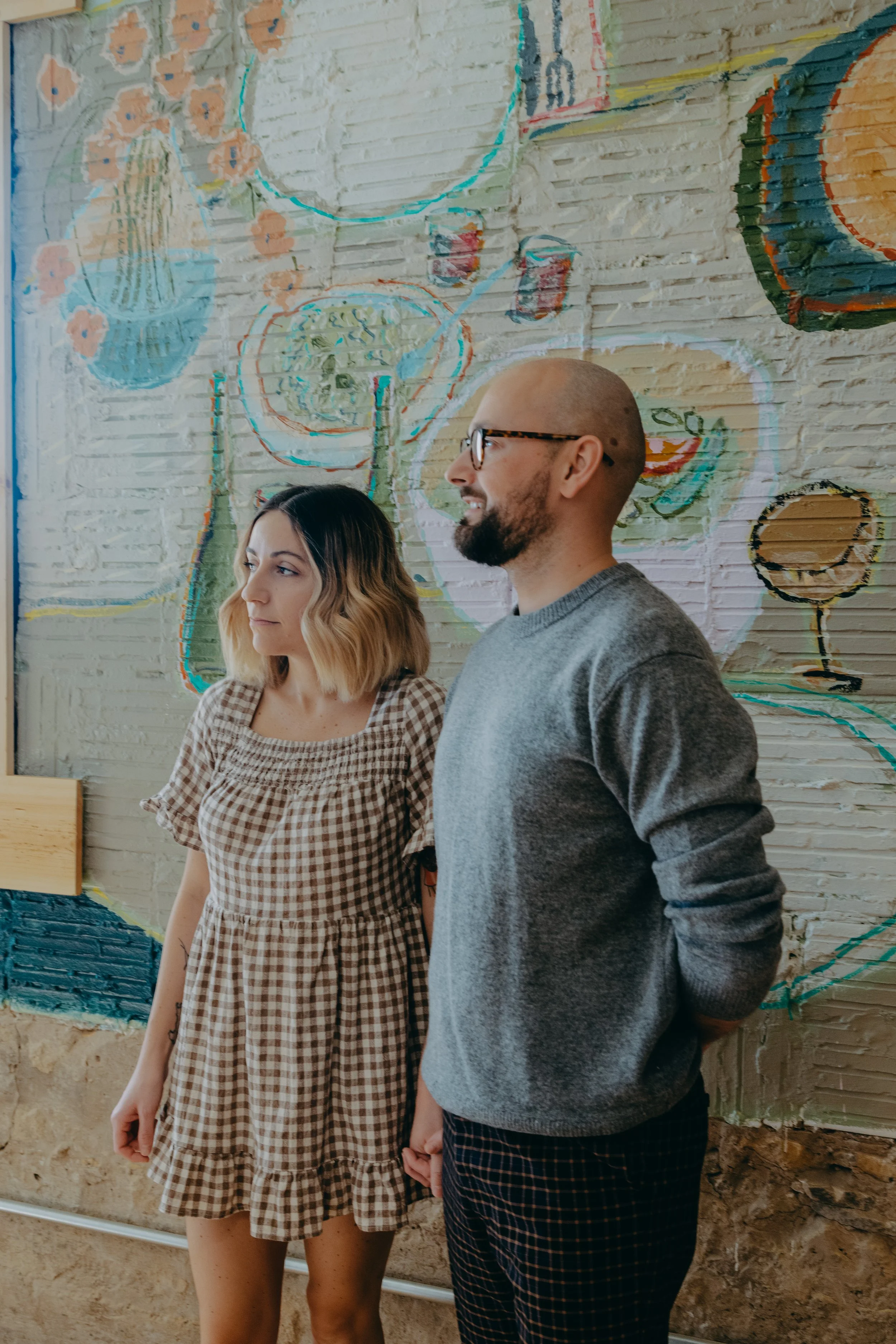 A woman and a man standing side by side in front of a colorful mural on a brick wall, holding hands.