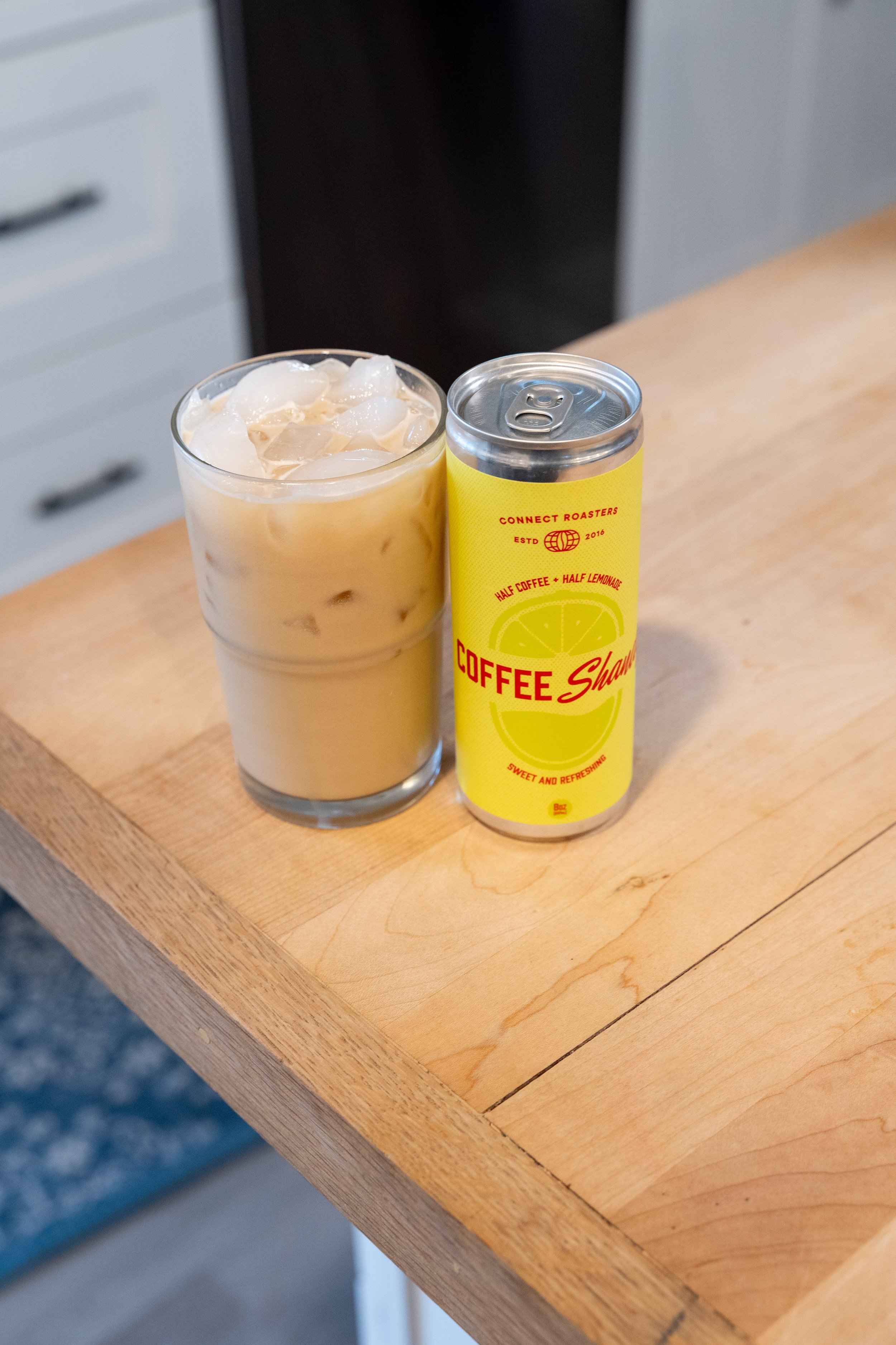 A glass of iced coffee next to a can of coffee lemonade on a wooden table.
