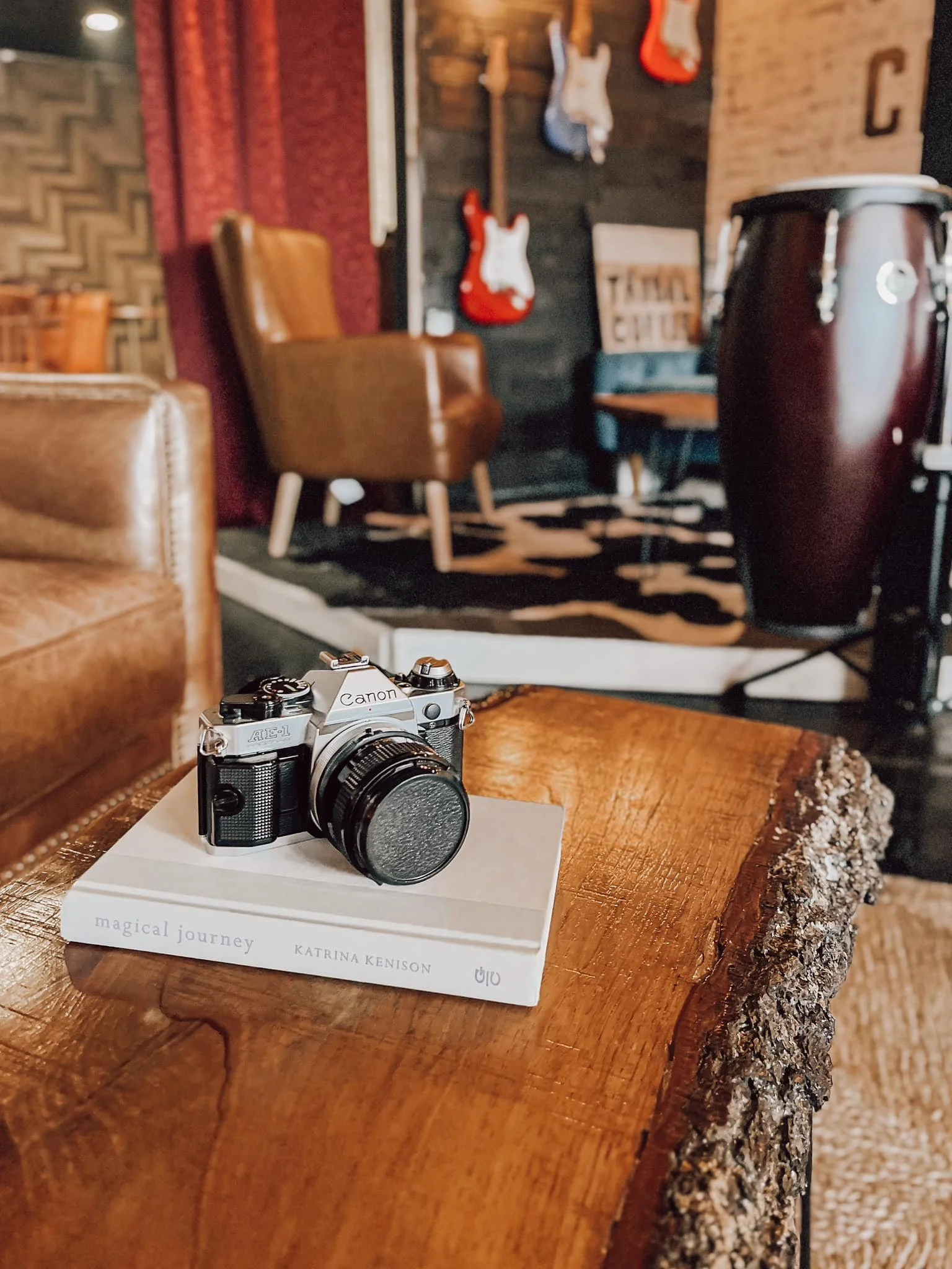 A vintage Canon AE-1 film camera placed on a white book titled 'magical journey' in a cozy room with leather furniture, guitars mounted on the wall, a large percussion instrument, and a patterned rug in the background.