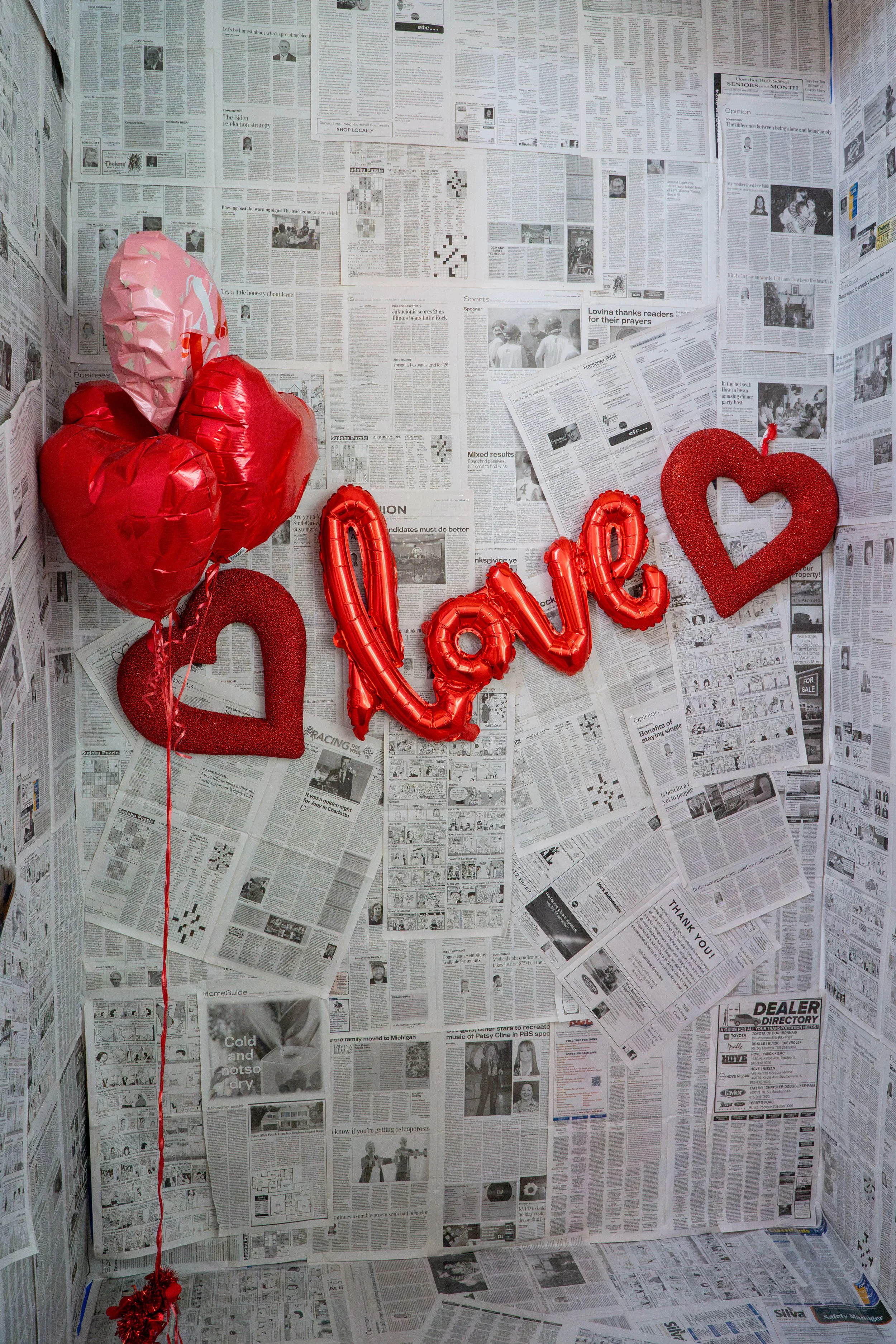 Valentine's Day decorations with red and pink balloons, glitter hearts, and a red 'love' balloon on a newspaper-covered background.