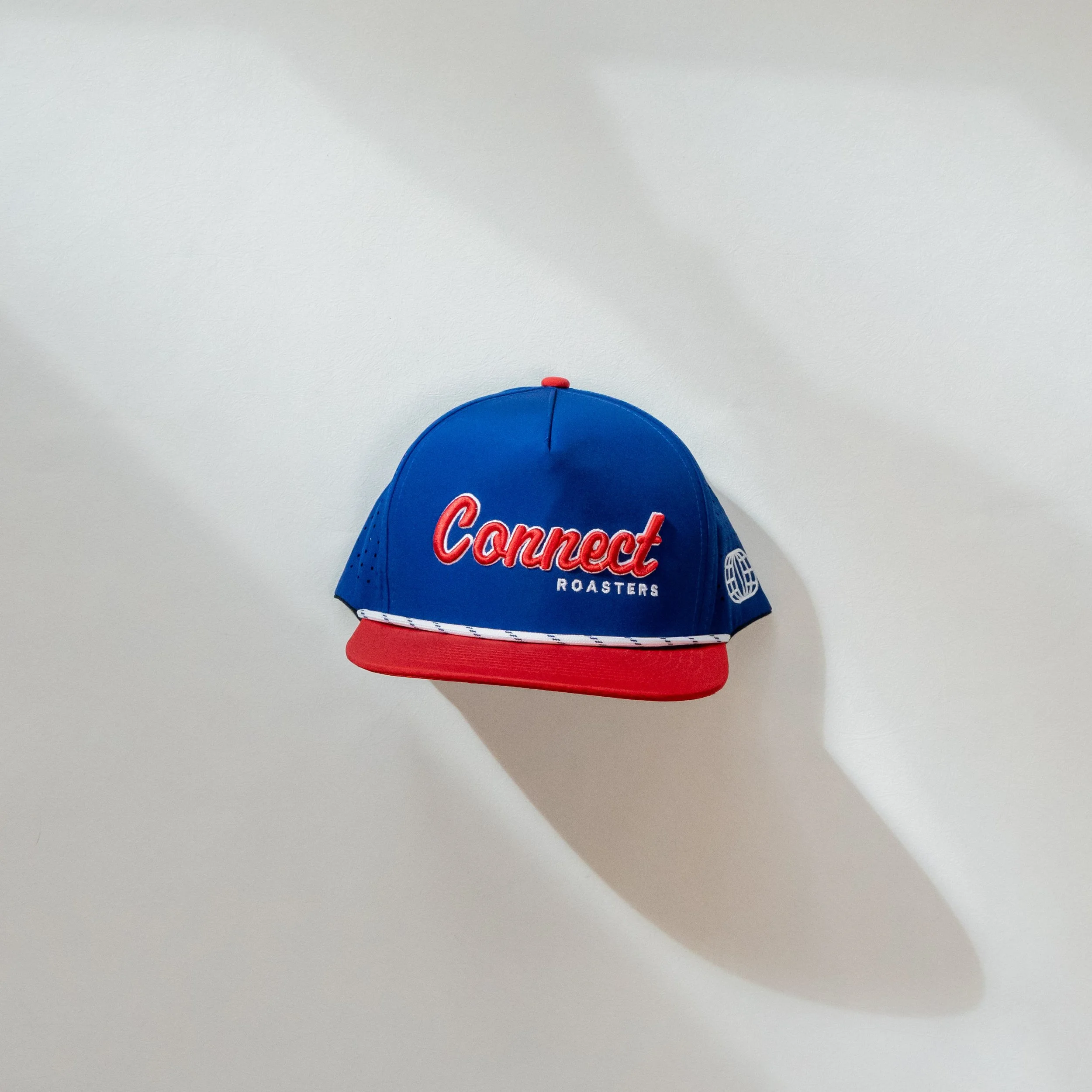 A blue and red baseball cap with the words 'Connect Roasters' embroidered on the front, placed on a white surface. Drop shadow to the right and below the cap.