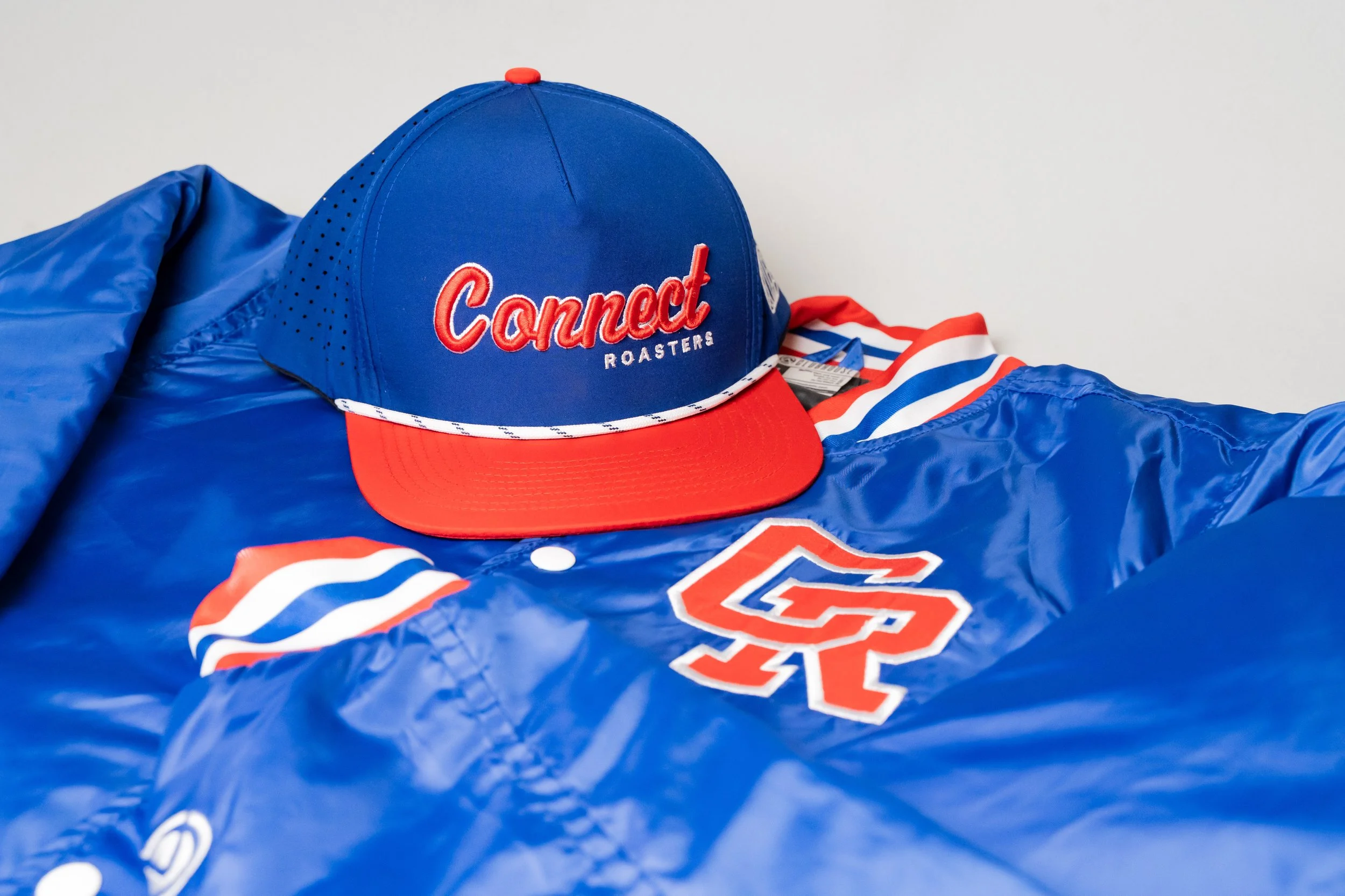 A blue baseball cap with red and white embroidery reading "Connect Roasters" atop a blue satin jacket with red, white, and blue striped details and a large embroidered "C" letter patch.