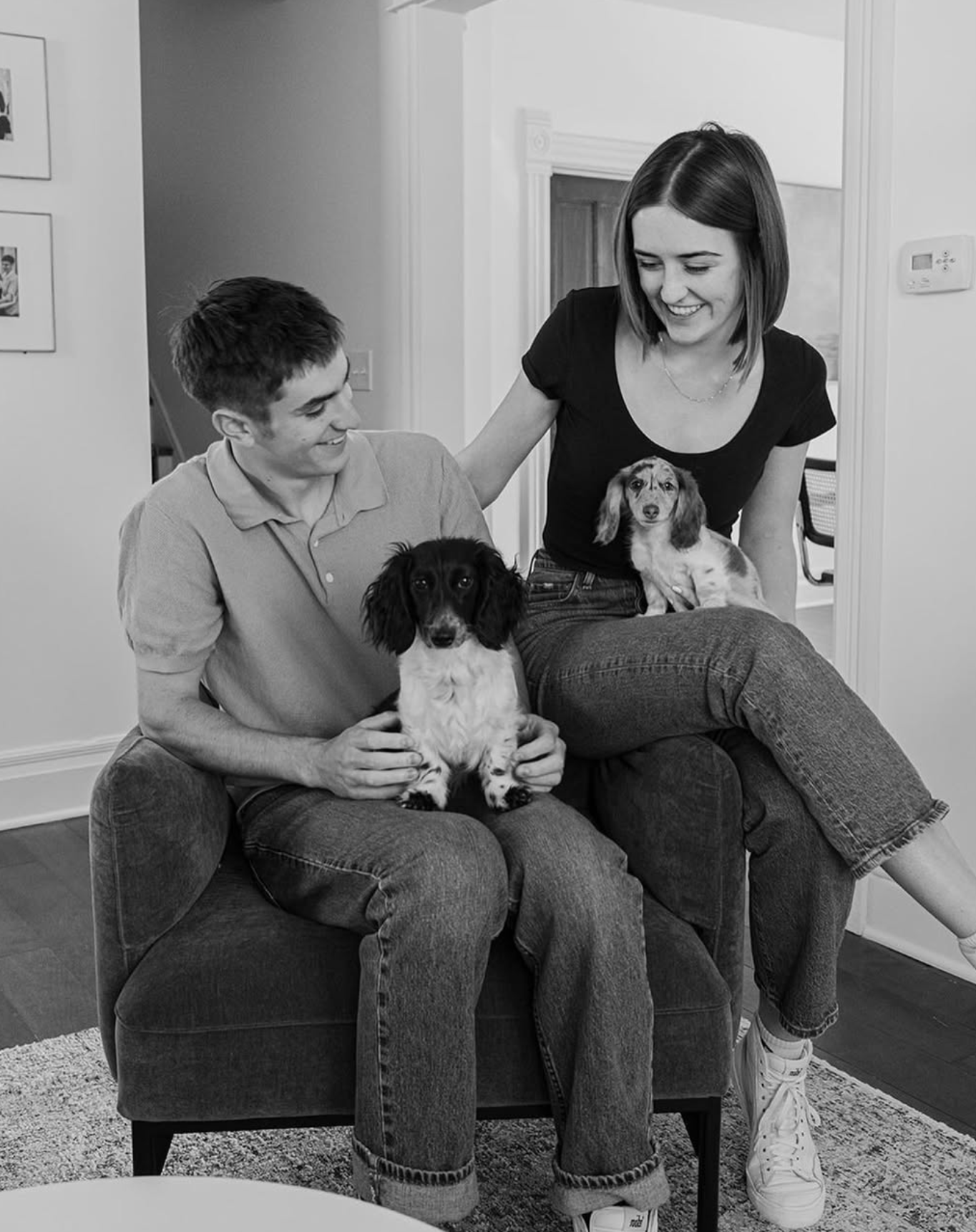 A smiling woman and a smiling man sitting on a couch with two cute dogs in a cozy living room.