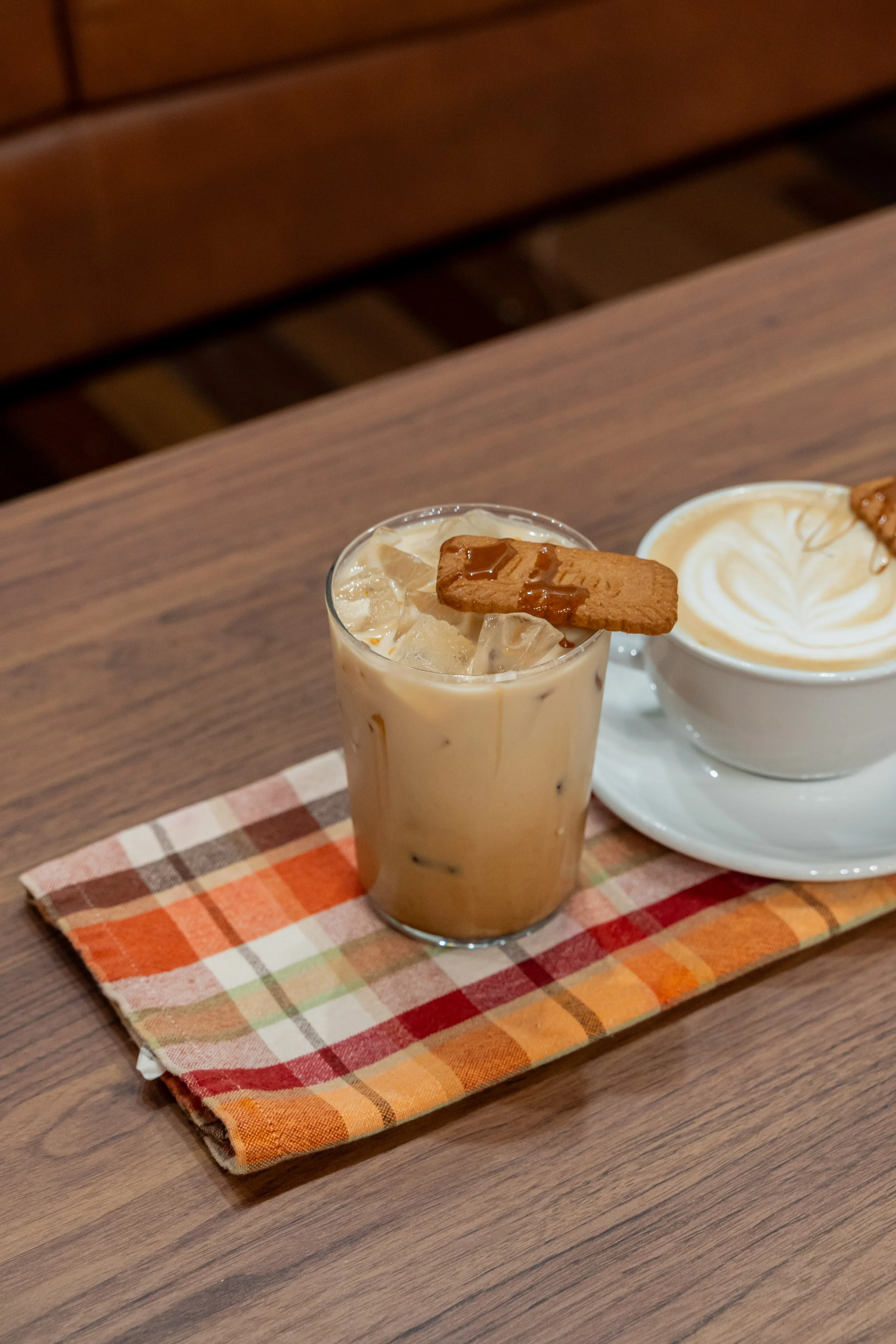 A glass of iced latte with a biscuit on top and a cup of latte art coffee on a saucer, placed on a checkered cloth on a wooden table.