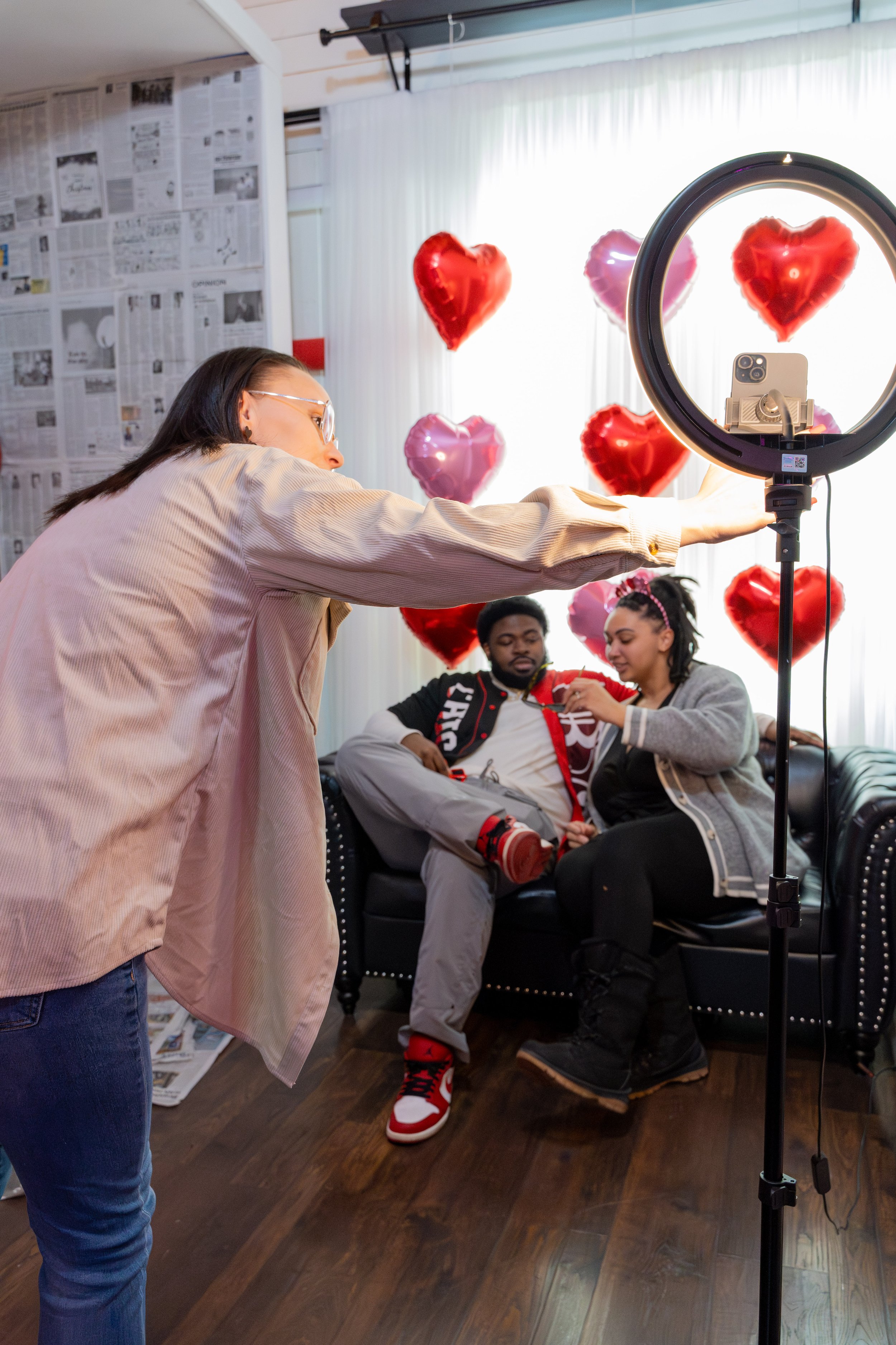 A woman takes a photo or records a video of a couple sitting on a black leather couch with heart-shaped balloons in the background. The man wears a black varsity jacket, gray pants, and red sneakers. The woman wears a gray jacket, black pants, and bl