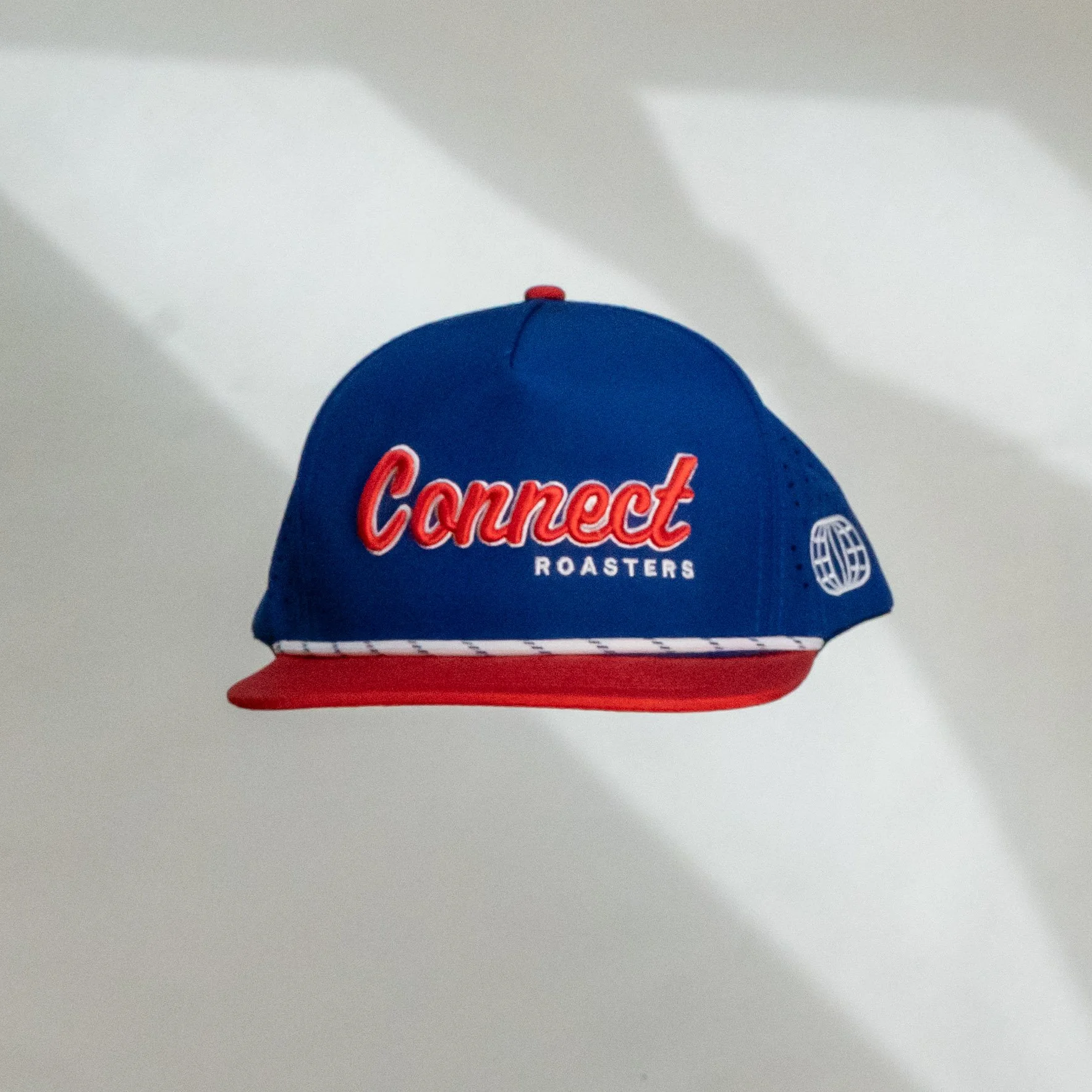 Blue and red baseball cap with 'Connect Roasters' embroidered on the front, hanging on a wall with shadows.