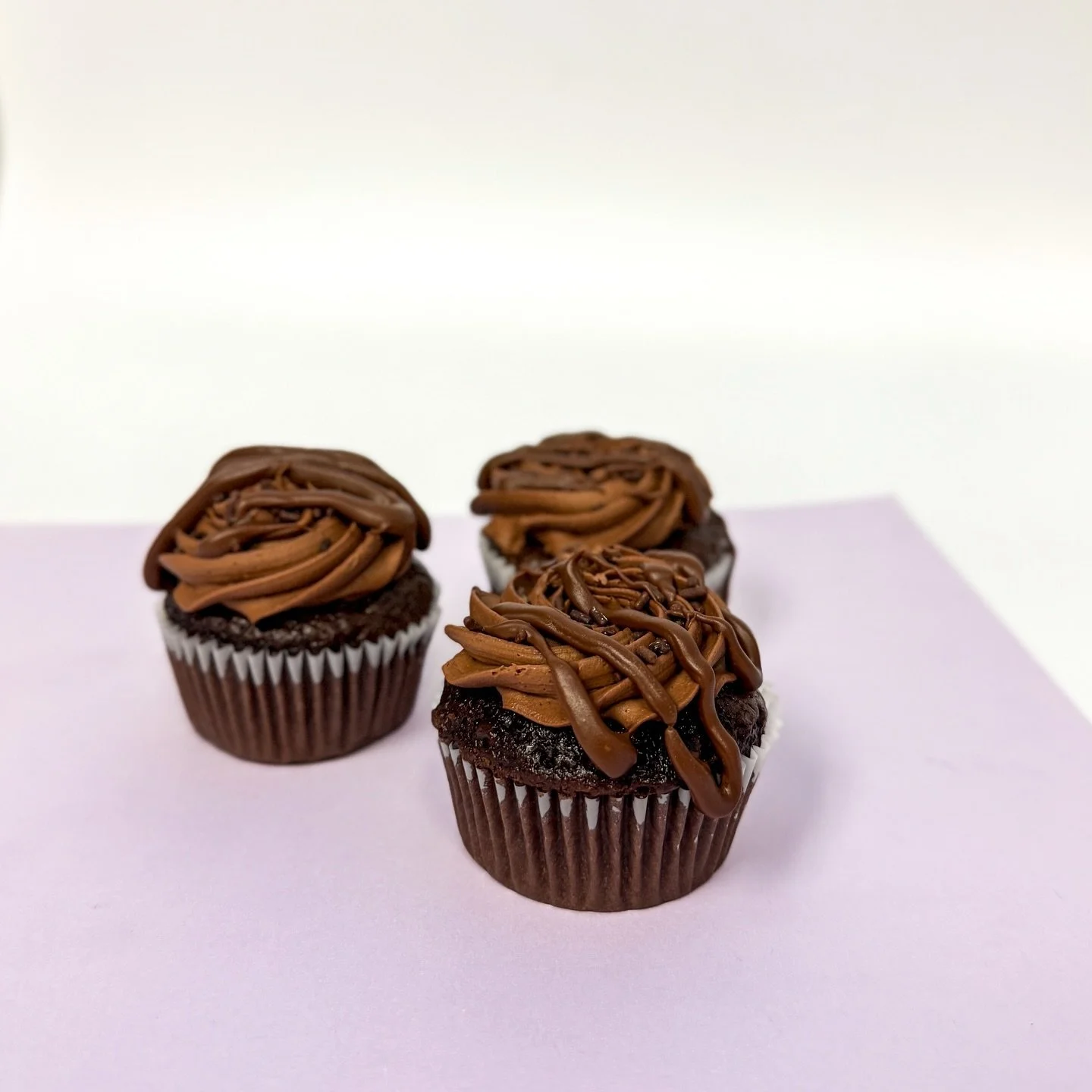 Three chocolate cupcakes with swirled chocolate frosting on top placed on a light purple surface.