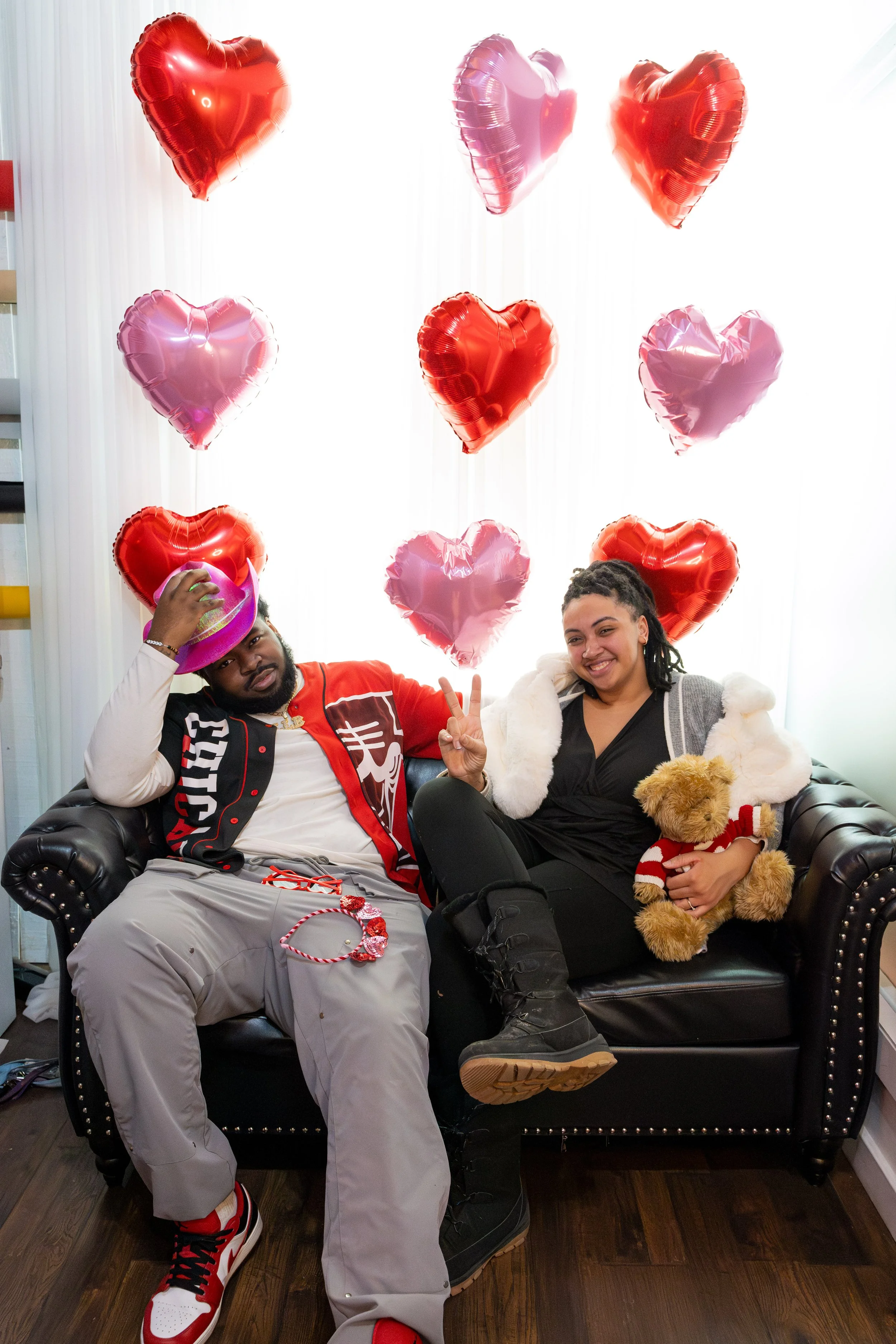 A man and woman sitting on a black leather couch with red and pink heart-shaped balloons hanging behind them. The man is wearing a red and black jacket, gray pants, and a pink hat, holding a pink hat in his hand. The woman is smiling, holding a teddy