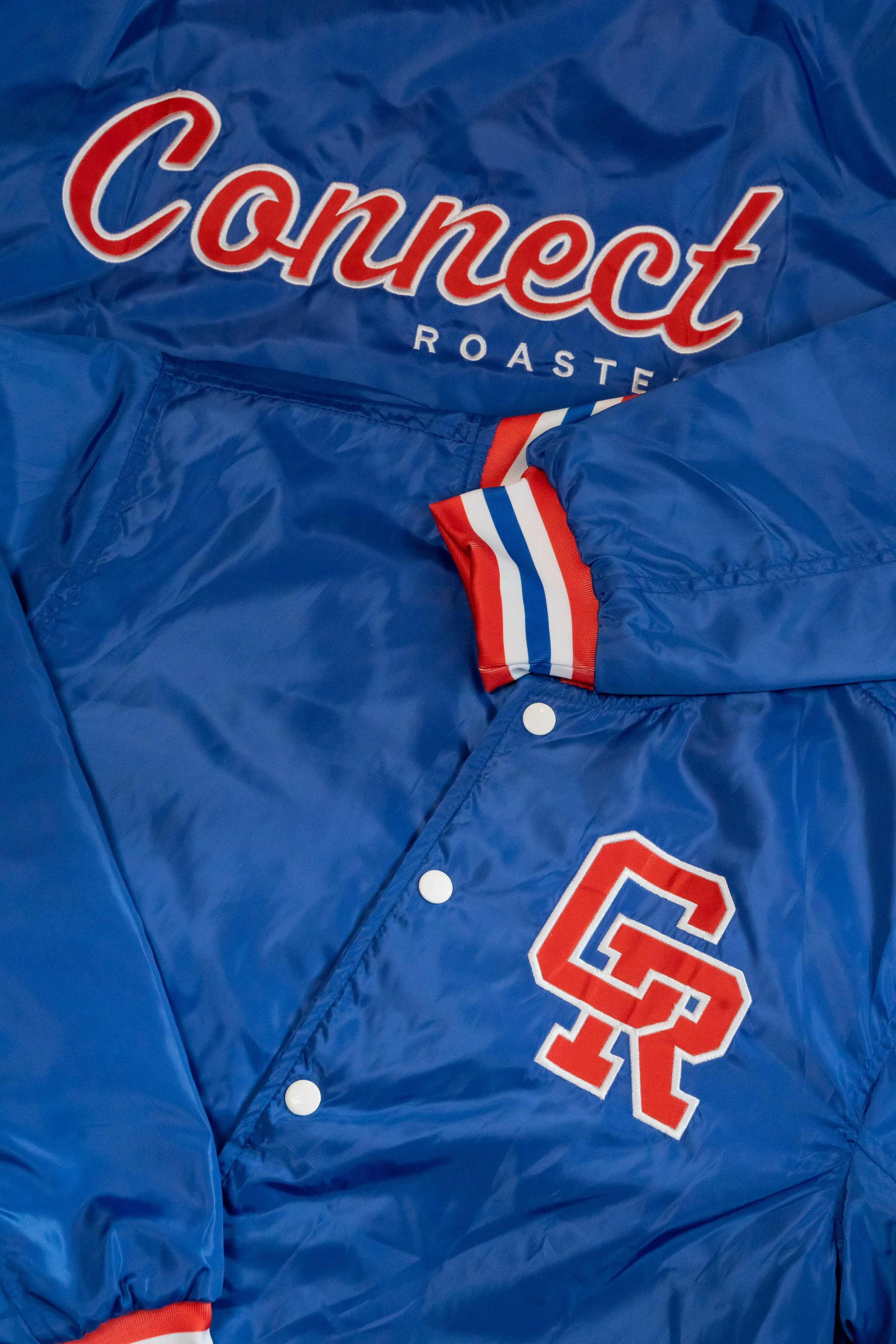 Close-up of a blue satin jacket featuring the text 'Connect Roaste' and a patch with the number 22, with red, white, and blue striped ribbing on the collar and cuffs.
