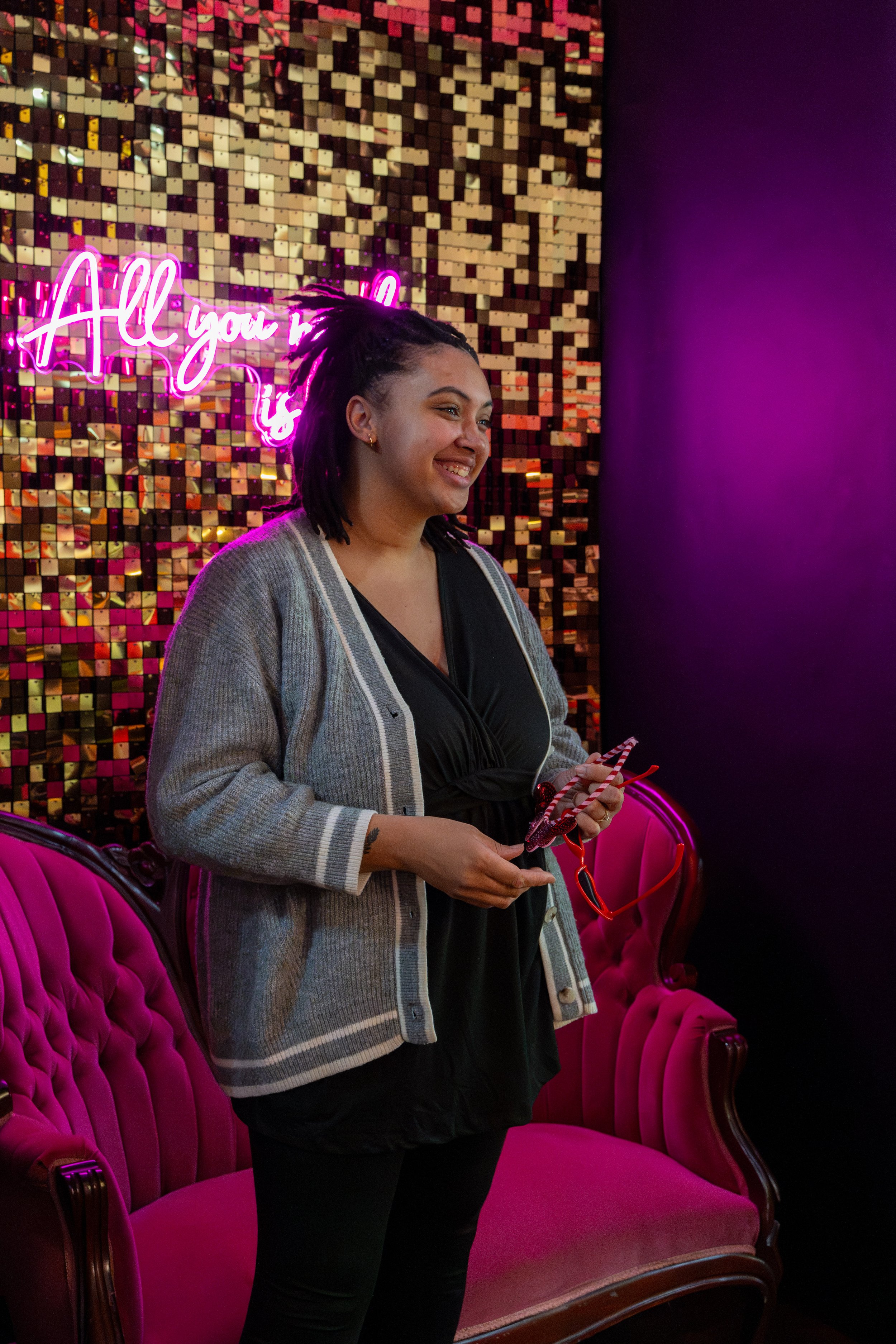 A woman with dreadlocks smiling, standing in front of a gold and pink mosaic wall with a neon sign that reads "All you need is love", holding sunglasses and a striped parasol, in a room with pink velvet furniture and purple lighting.