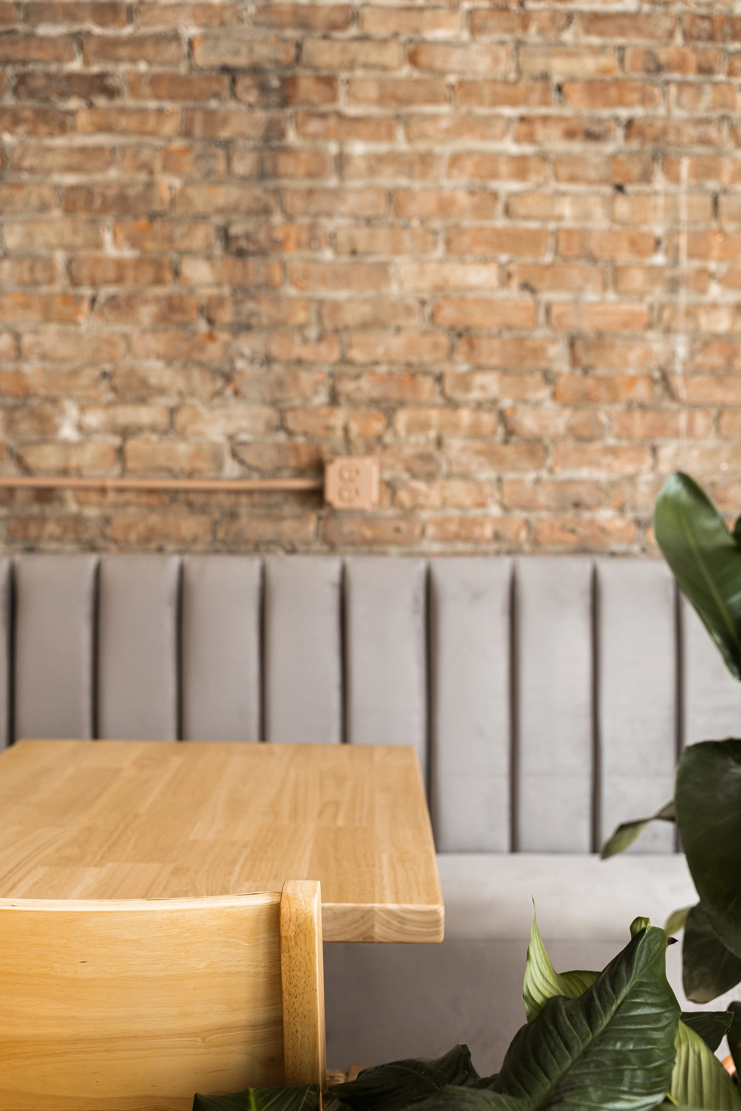 Interior view of a minimalist café with a wooden table, a beige upholstered bench, a gray wall, and a exposed brick wall background. A green plant is partially visible in the foreground.