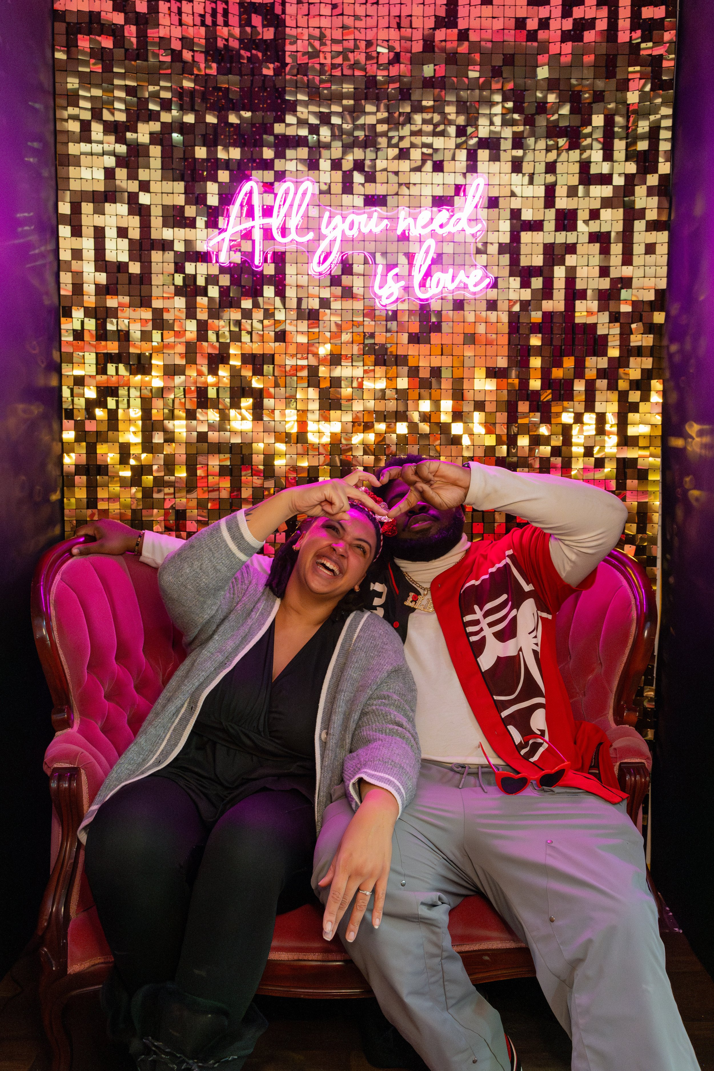 Two people sitting on a pink vintage sofa, making a heart shape with their hands in front of a golden mosaic wall with a pink neon sign that reads 'All you need is love'.