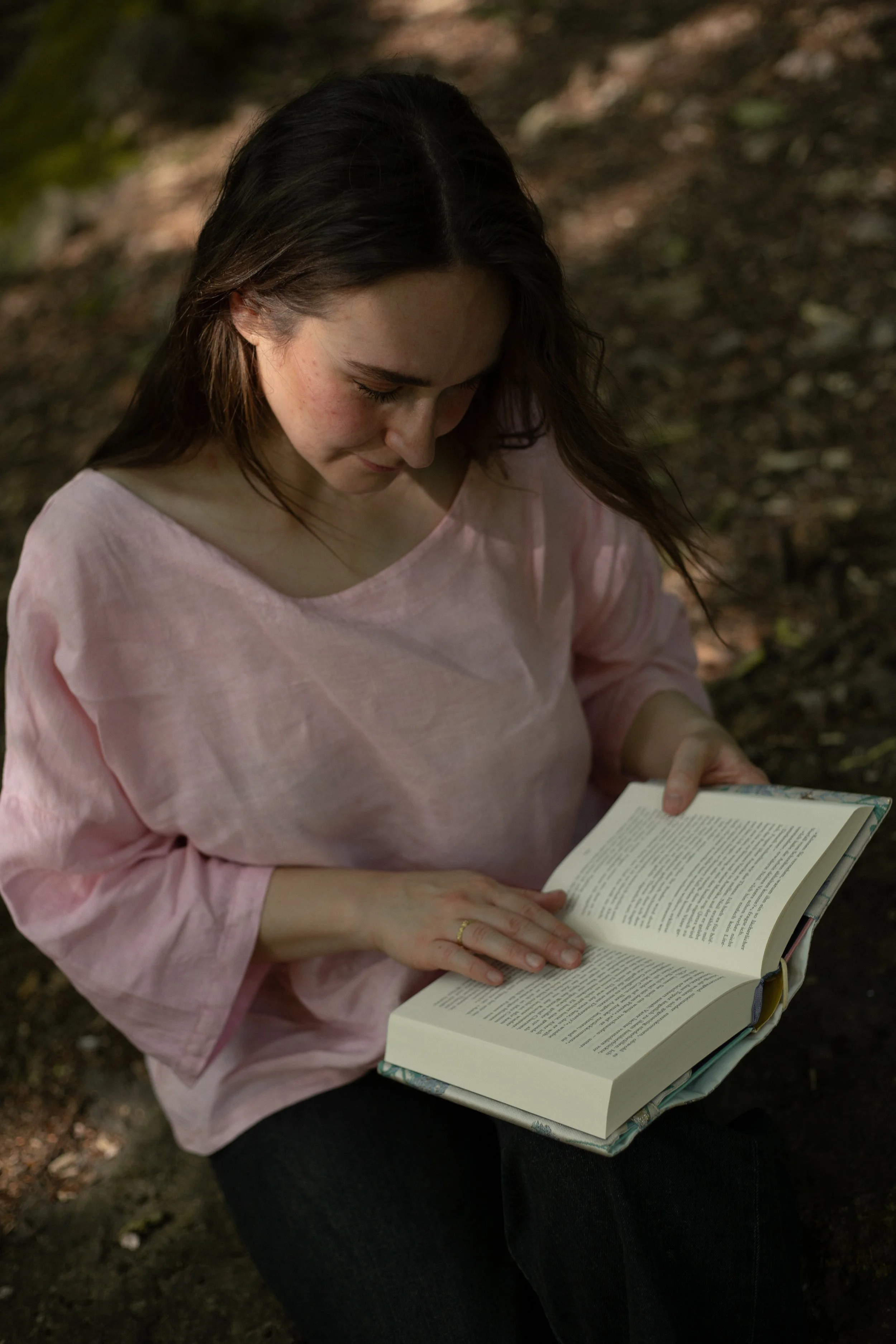 Lektorin Monique Reinbold sitzt vor einem verschwommenen braunen Hintergrund in der Natur. Sie hat lange braune Haare und trägt ein rosa Leinenshirt. In ihren Händen hält sie ein geöffnetes Buch, auf welches sie ihren Blick richtet.