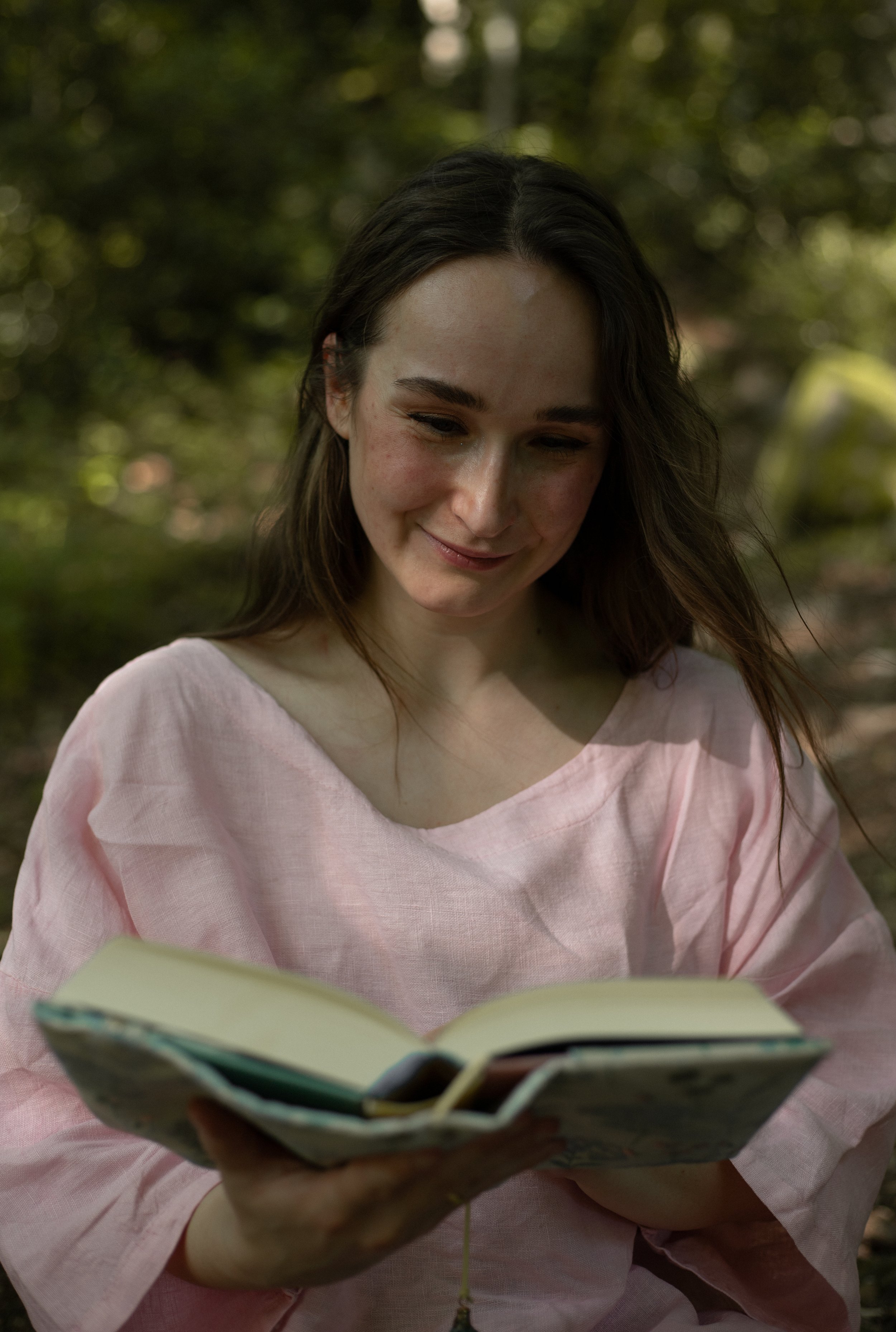 Lektorin Monique Reinbold ist vor einem verschwommenen grünen Hintergrund zu sehen. Sie hält ein offenes Buch in der Hand, auf welches sie ihren Blick richtet.