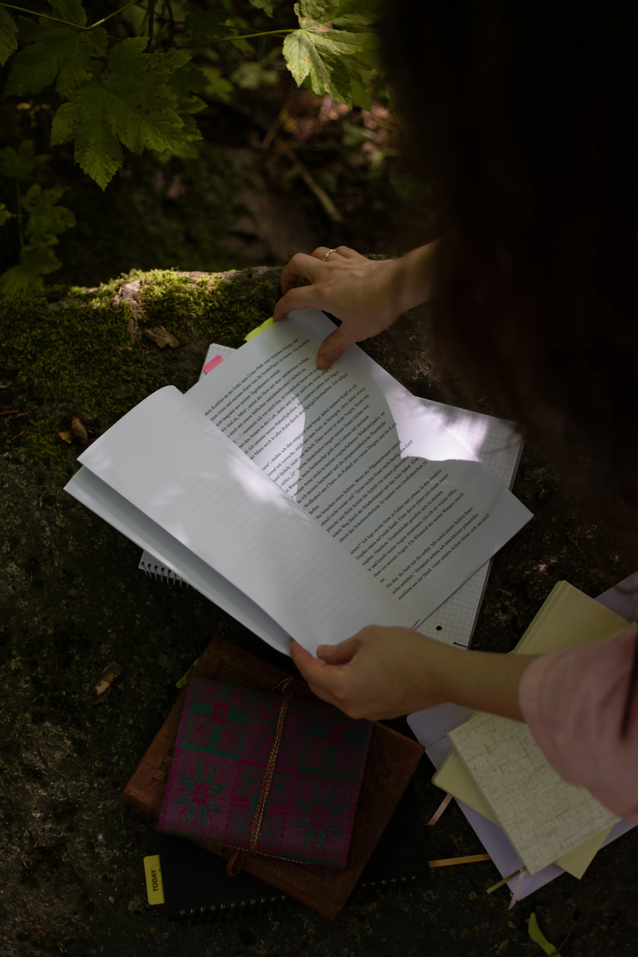 Auf einem moosigen Stein liegt ein Manuskript, welches mit gelben und pinken Post-Its markiert wurde. Hände blättern durch dessen Seiten.