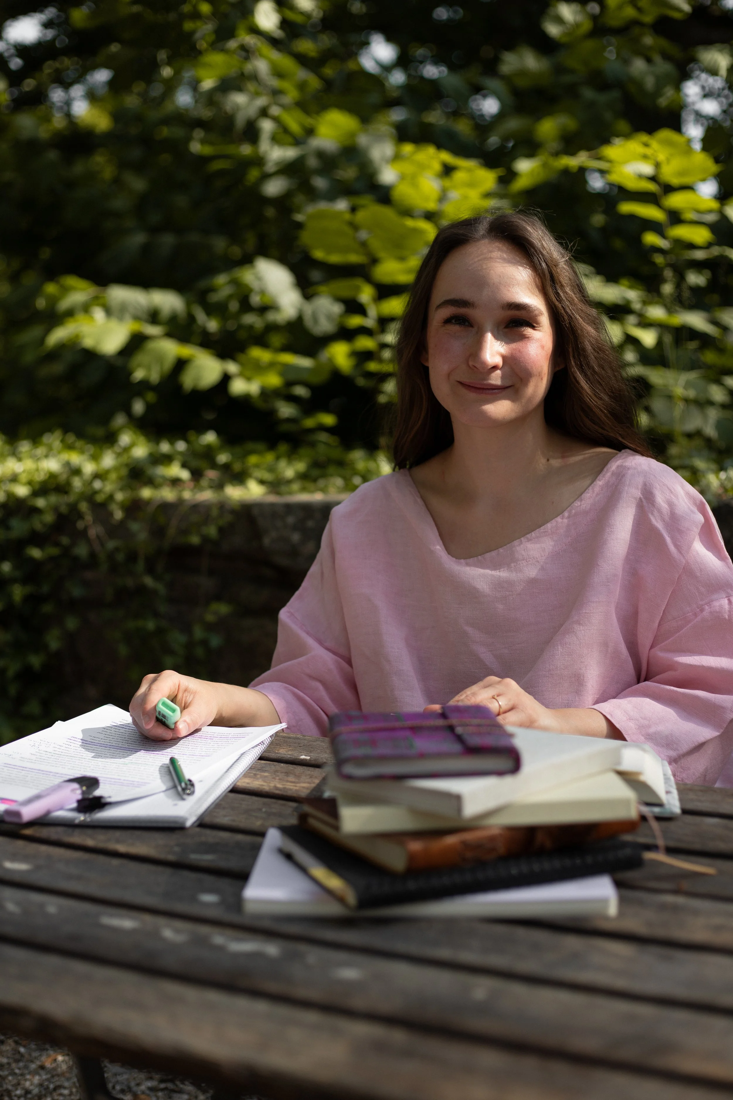 Lektorin Monique Reinbold von Lektorat Komerebi sitzt an einem Holztisch in der Natur. Sie blickt mit einem freundlichen Gesichtsausdruck in die Kamera. Sie trägt ein rosa T-Shirt aus Leinen. Vor ihr liegen gestapelte Bücher und ein Manuskript.