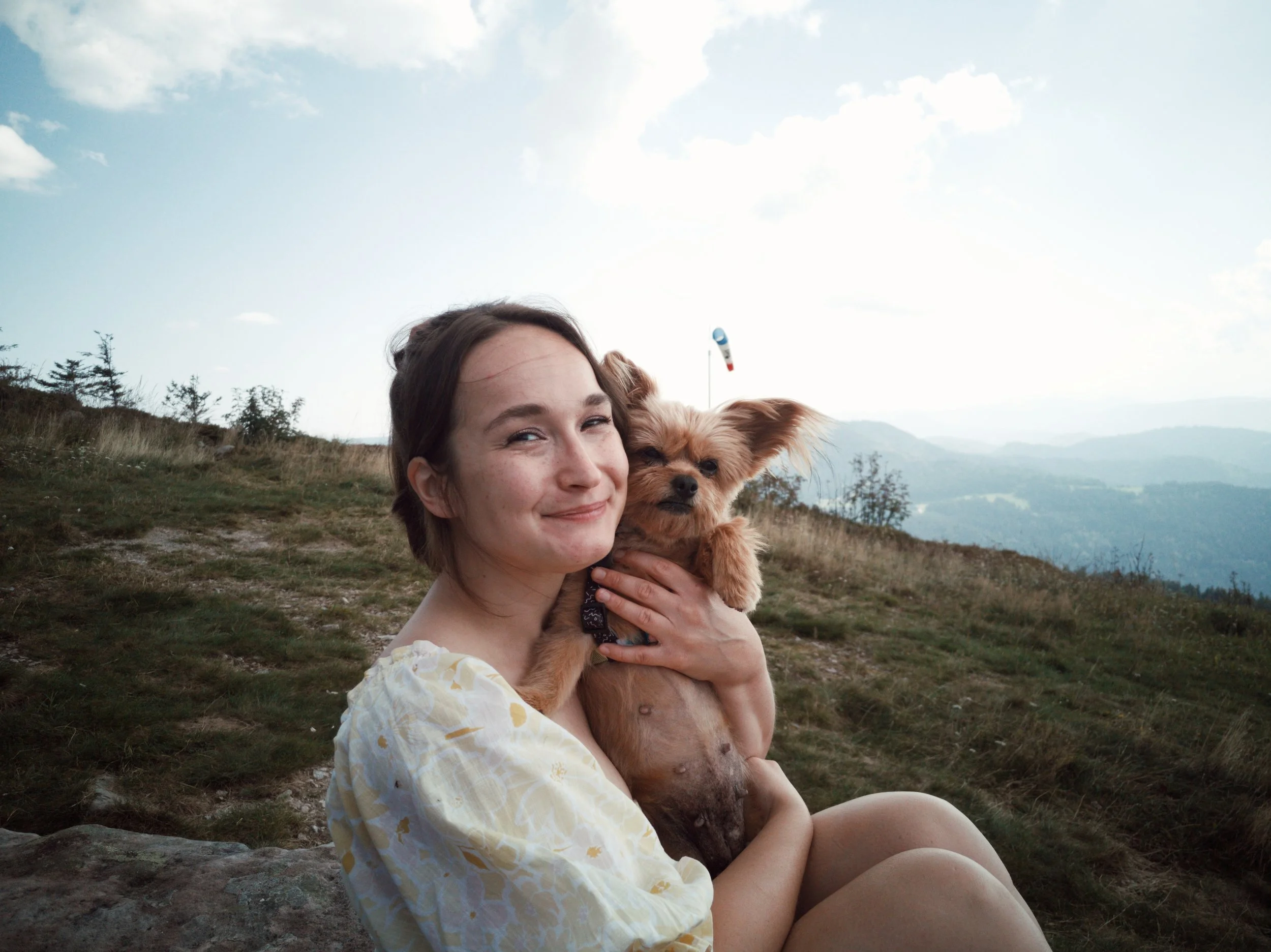 Lektorin Monique Reinbold sitzt auf einer Bergwiese. Ihre braunen Haare trägt sie in einem Zopf. Sie lächelt in die Kamera. In ihren Armen hält sie einen kleinen braunen Hund mit großen Ohren und wuscheligem Fell.