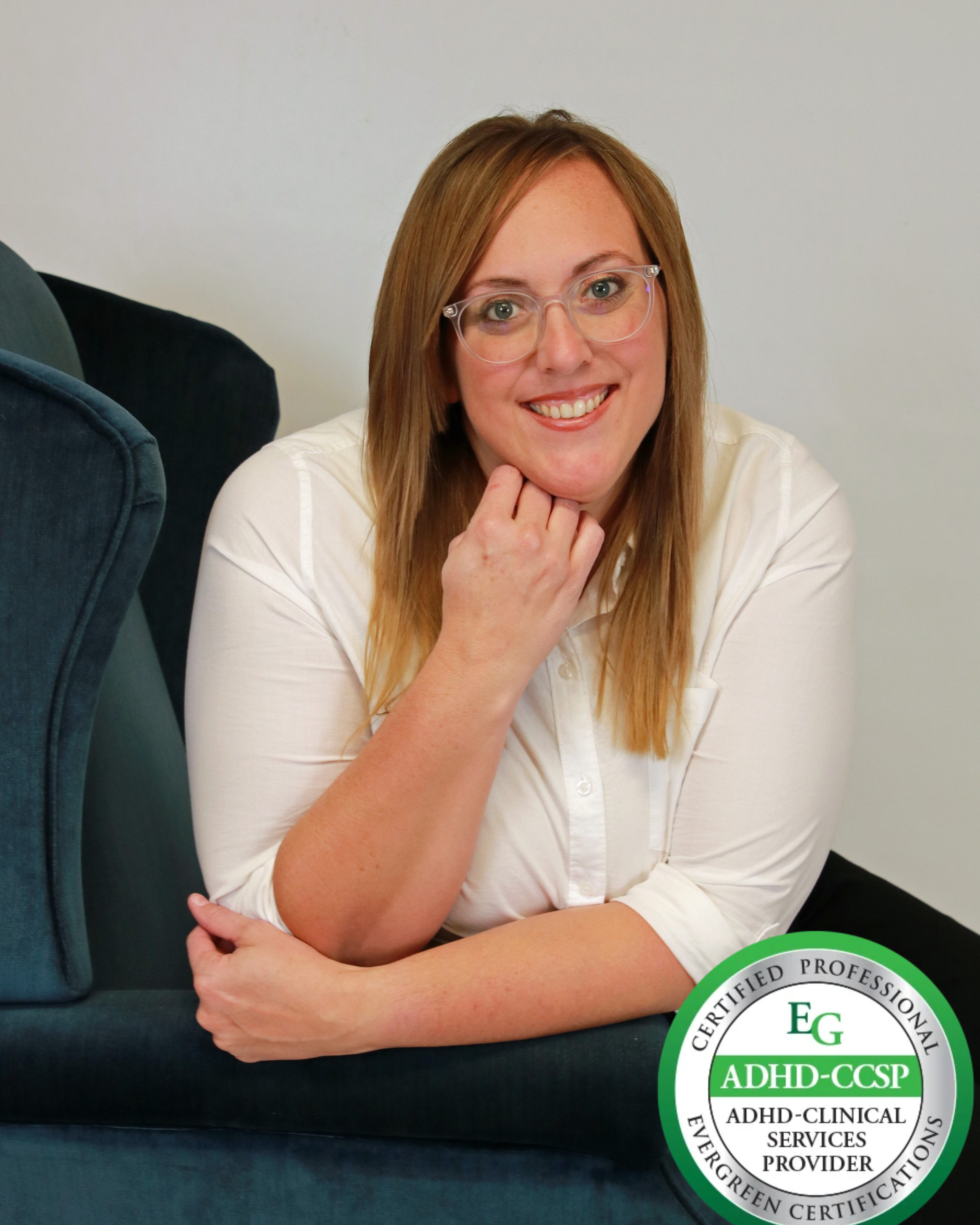 A woman with glasses and long red hair, smiling, sitting next to a teal-colored armchair against a plain white background.