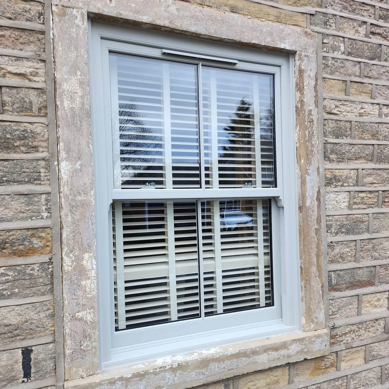 A white-framed double-hung window with security shutters on a brick house. The window reflects trees and a car outside.