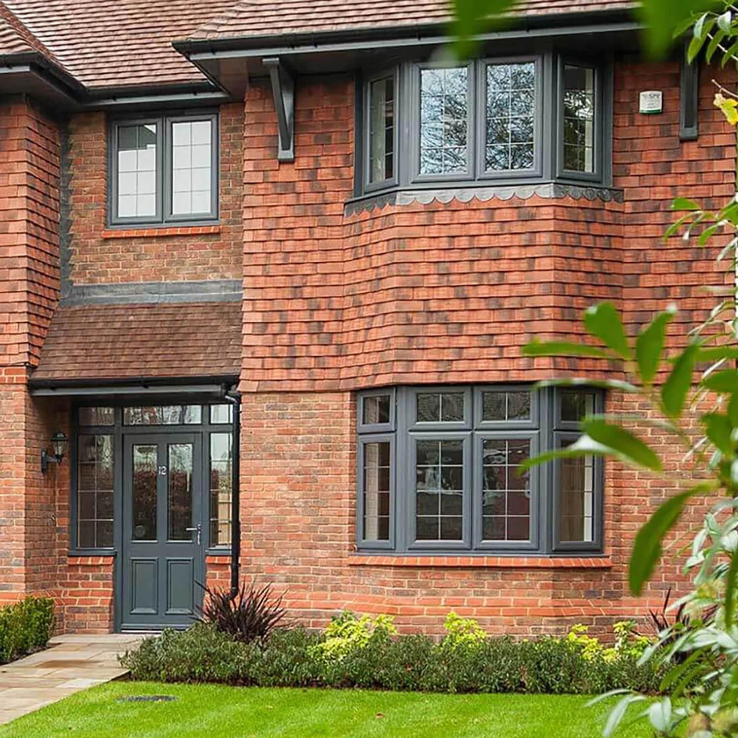 A brick house with multiple windows, including a bay window on the second floor, with black trims and a front door, surrounded by a well-maintained lawn and garden.