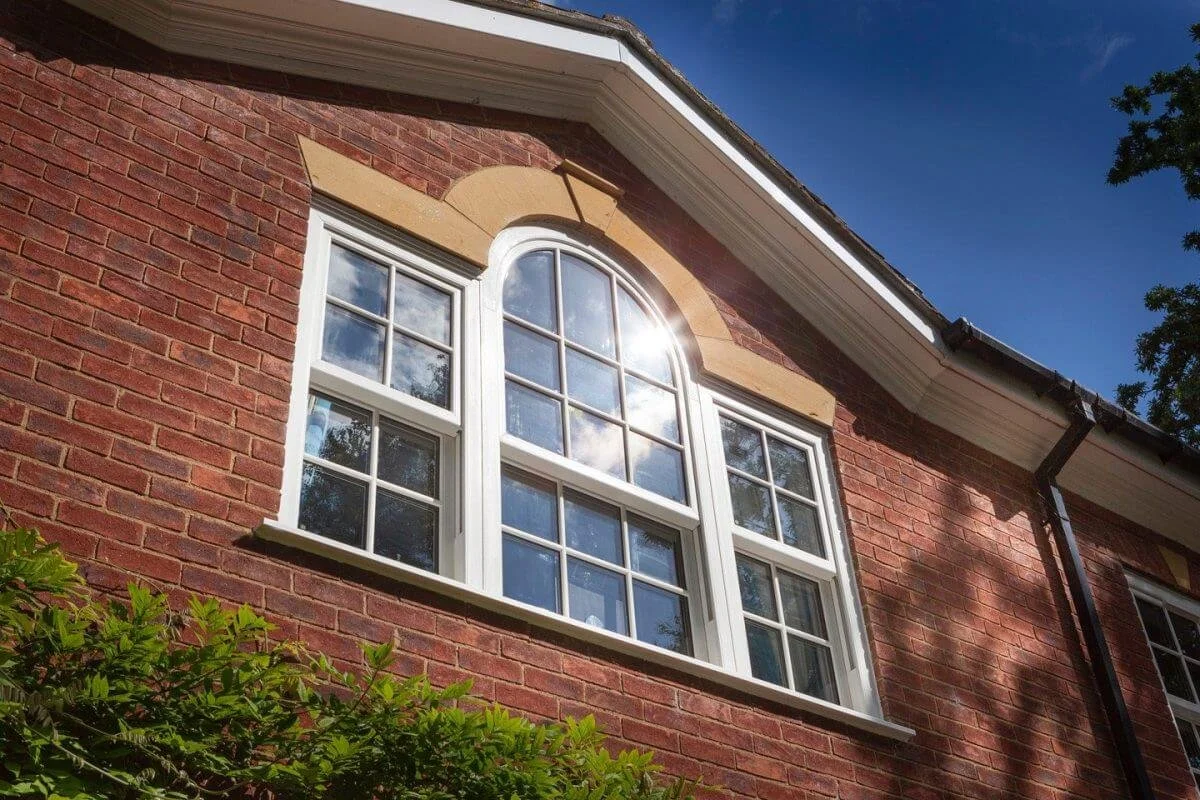 Close-up of a red brick house window with white framing and an arched top in bright sunlight, with trees and a blue sky reflected in the glass.